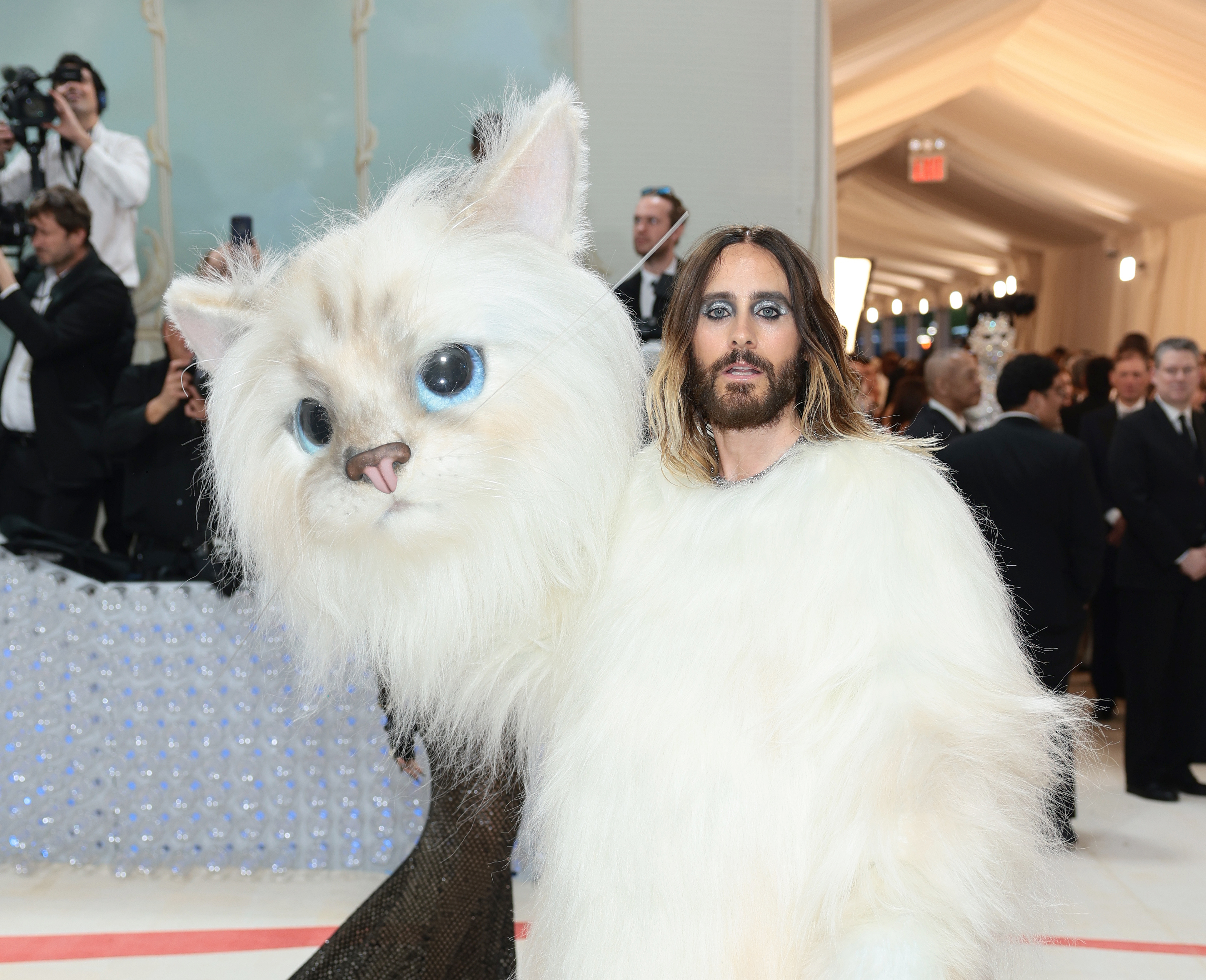 Jared Leto dressed in a white fluffy outfit holding a large cat head on the red carpet at an event, surrounded by photographers and attendees