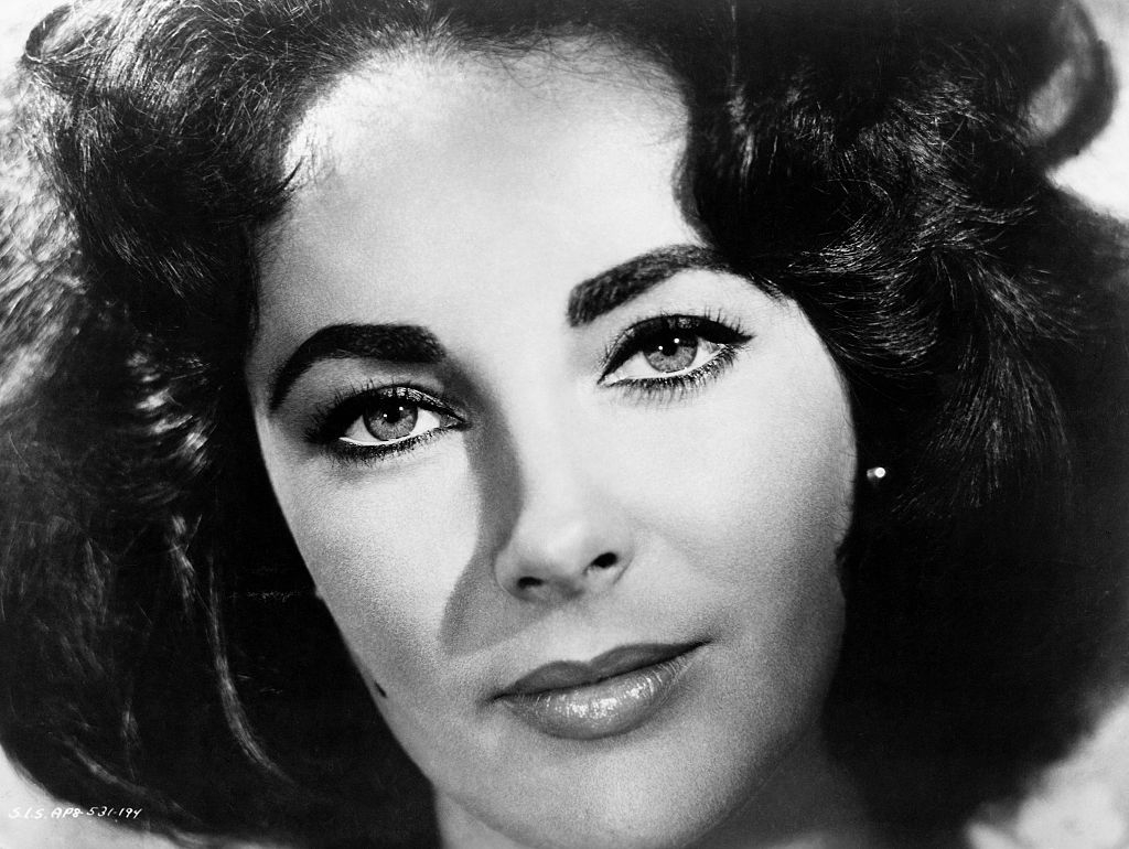 Close-up black and white portrait of Elizabeth Taylor with styled dark hair, strong eyebrows, and a subtle smile
