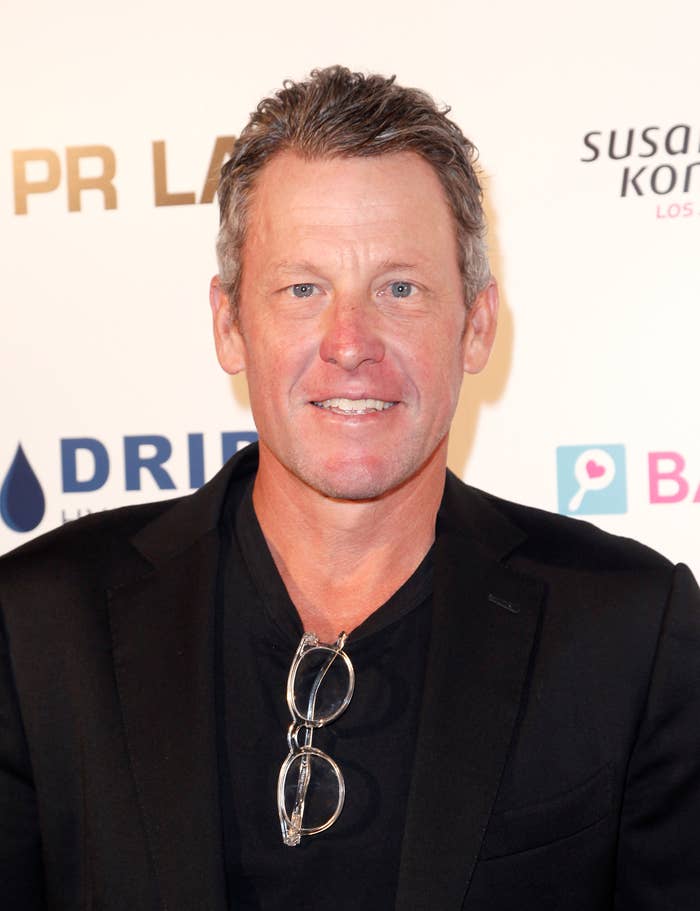 Lance Armstrong at a red carpet event wearing a black blazer and shirt, with glasses resting on his chest