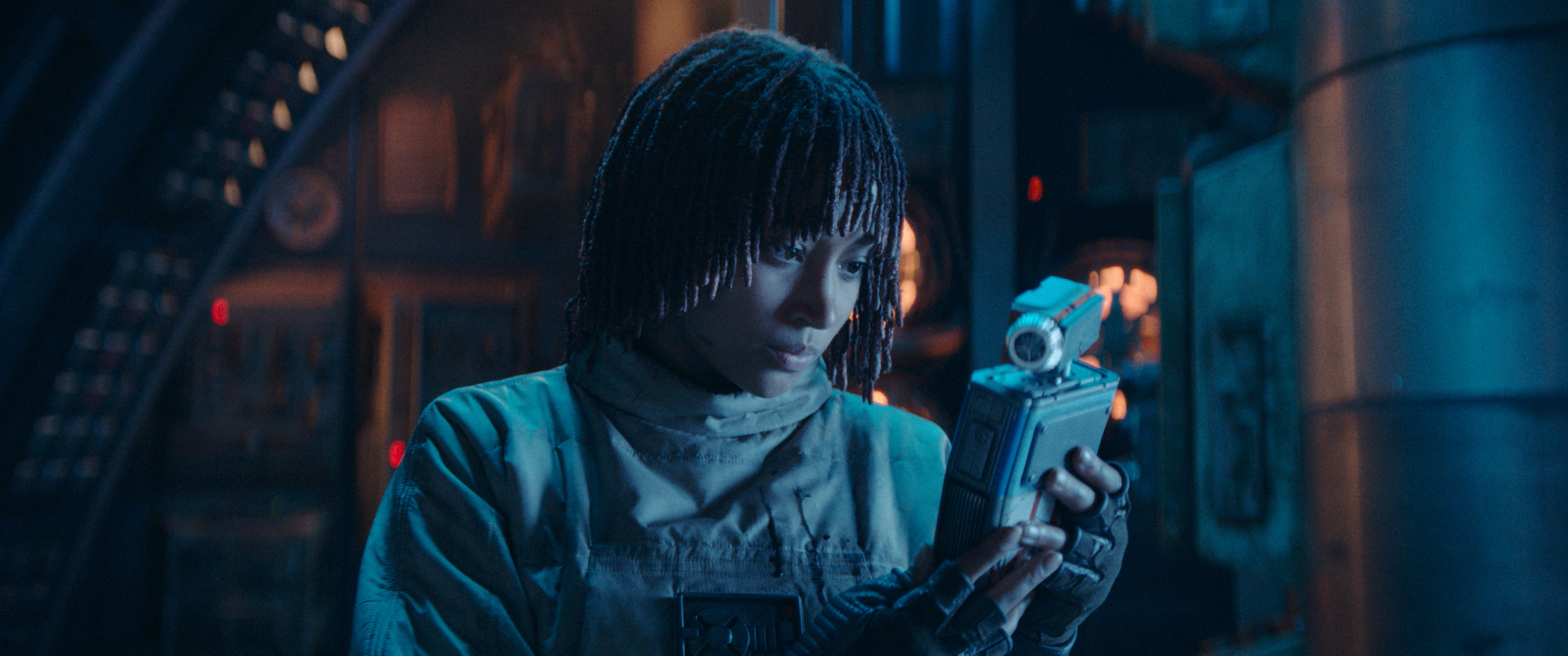 Amandla, dressed in a futuristic outfit, holds a gadget inside a high-tech, sci-fi setting, appearing focused and contemplative