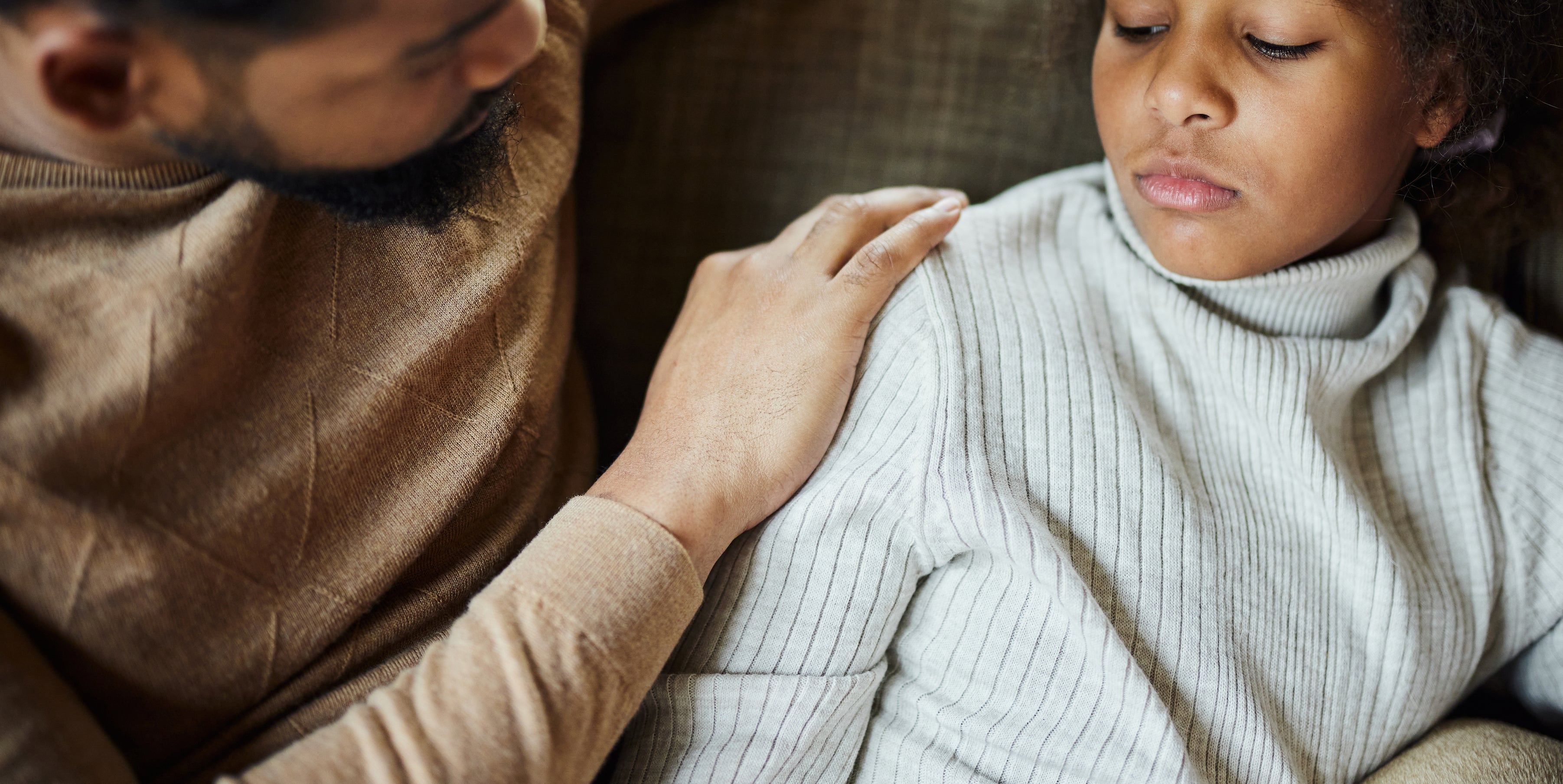A man with his arm around a sad or thoughtful girl, looking at her with concern as they sit on a couch