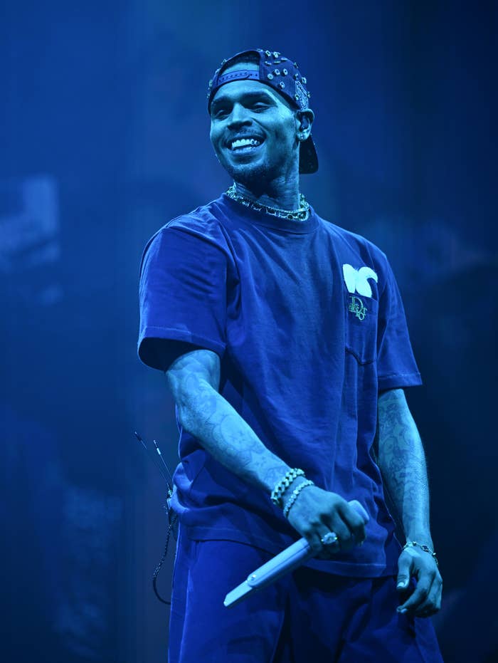 Chris Brown performing on stage, wearing a casual T-shirt, baseball cap, and accessories, smiling and holding a microphone