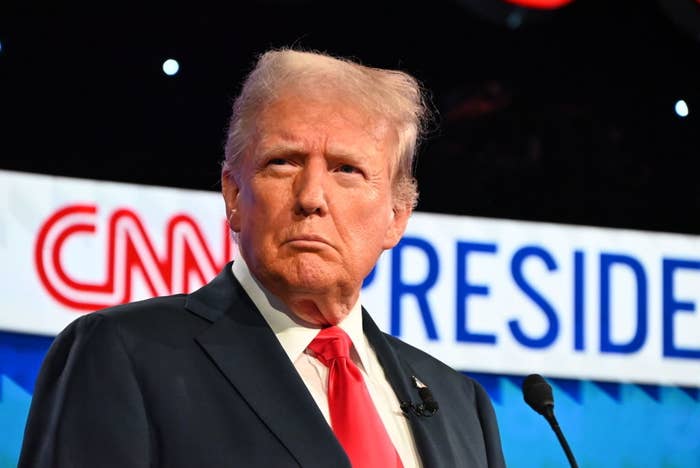 Donald Trump stands in front of a CNN sign that partially reads "President," wearing a suit and tie