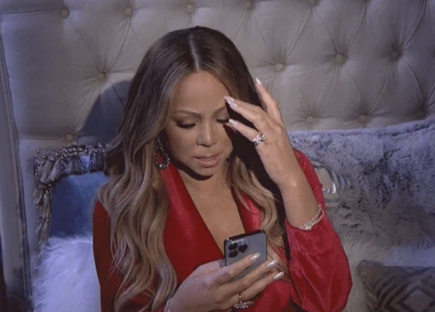 Mariah Carey in a red, silky outfit looking concerned while holding a smartphone and touching her forehead