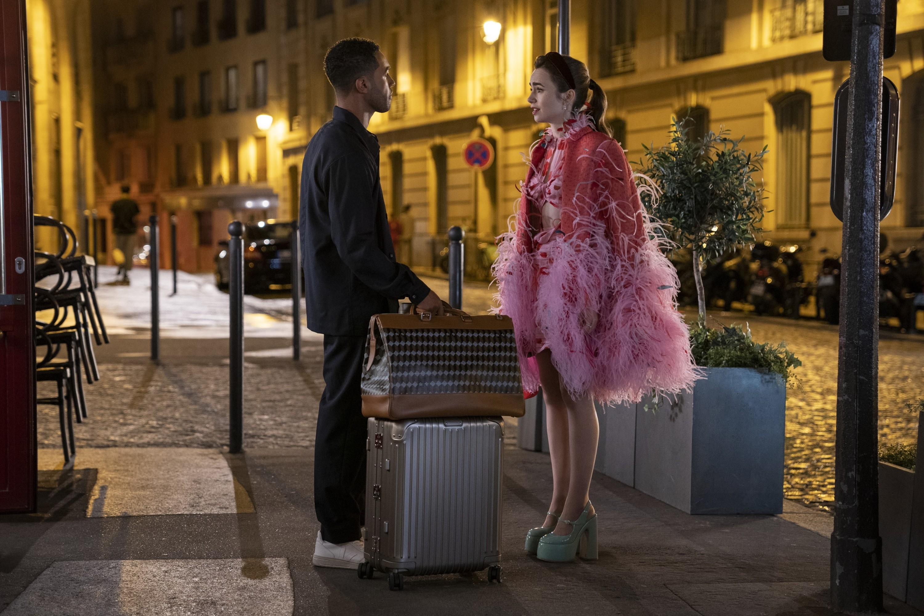 Lucien Laviscount and Lily Collins have a conversation in "Emily in Paris." He wears a dark outfit and she is wearing a pink feather cape and heels.