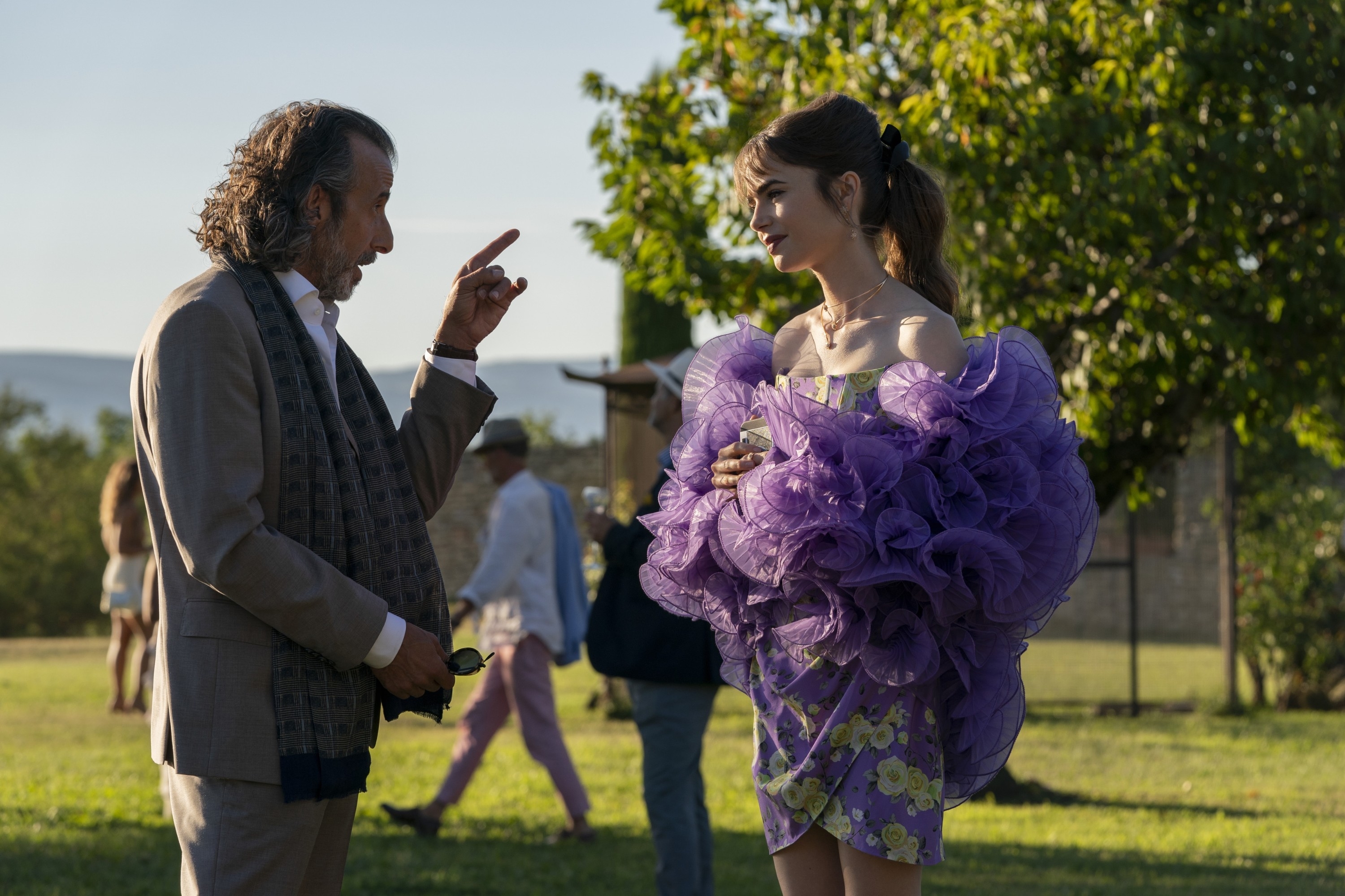 David Coburn and Lily Collins in a purple dress with ruffles and florals talking outdoors in "Emily in Paris."