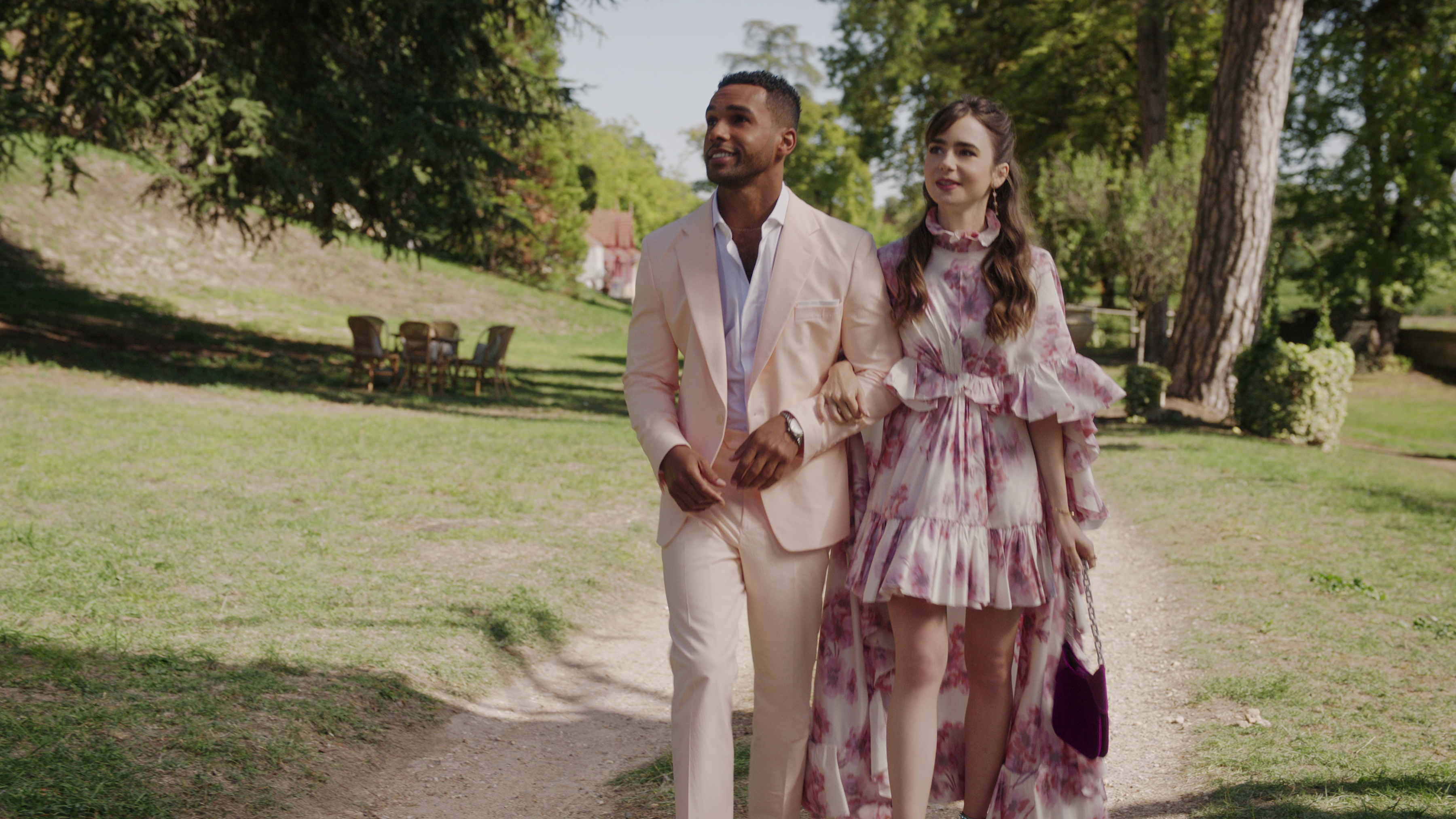 Lucien Laviscount and Lily Collins walk hand-in-hand outdoors. Lucien wears a suit, and Lily dons a floral, ruffled dress in "Emily in Paris."