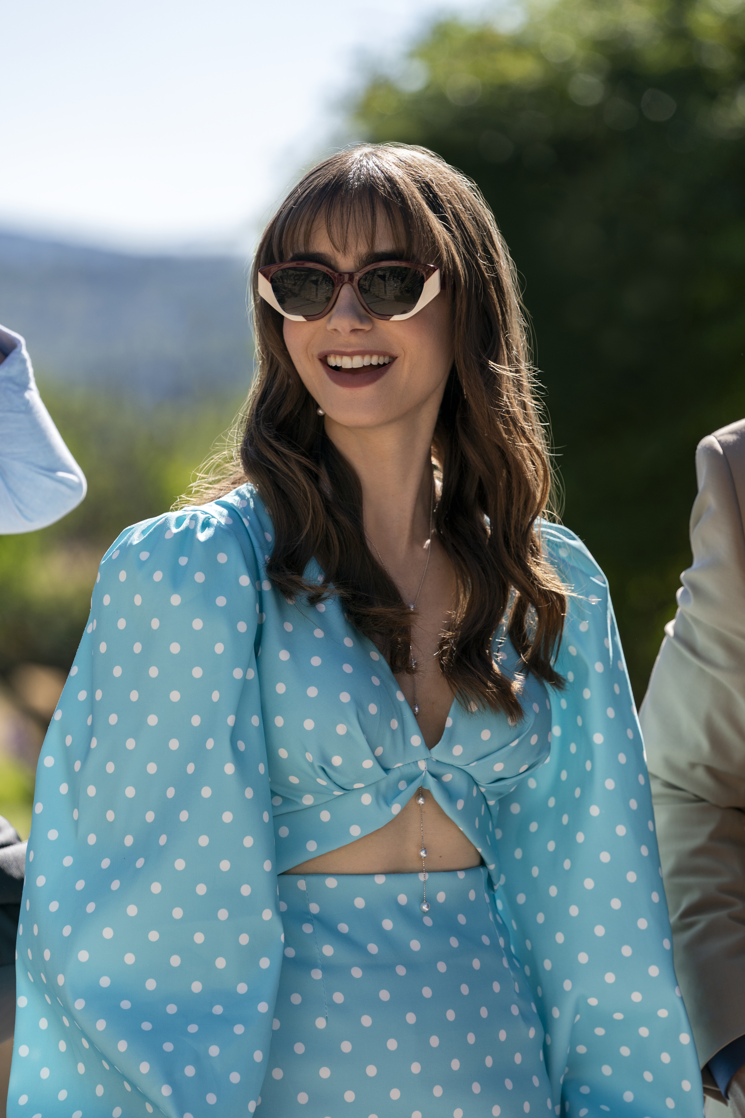 Lily Collins, wearing a stylish polka-dot dress with unique cutouts and sunglasses, smiles outdoors in "Emily in Paris."