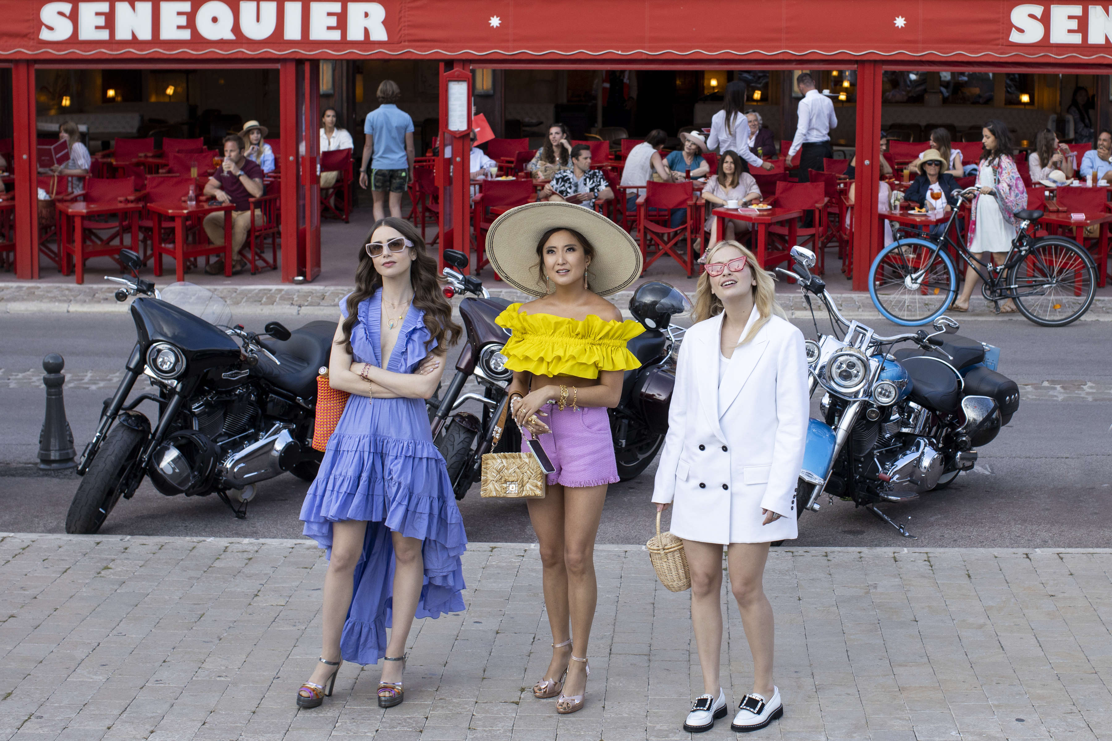 Lily Collins, Ashley Park, and Camille Razat stand in front of motorcycles in stylish outfits during a scene from "Emily in Paris."