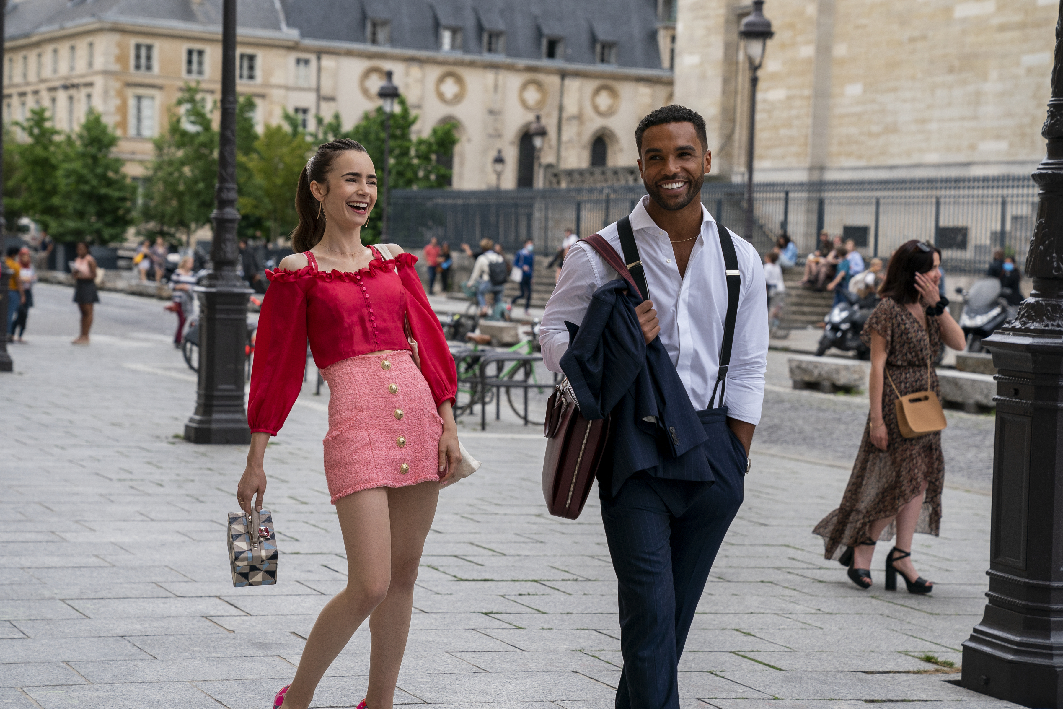 Lily Collins and Lucien Laviscount walk through a city square. Lily wears a red top with a pink skirt, while Lucien wears a white shirt with suspenders in "Emily in Paris."