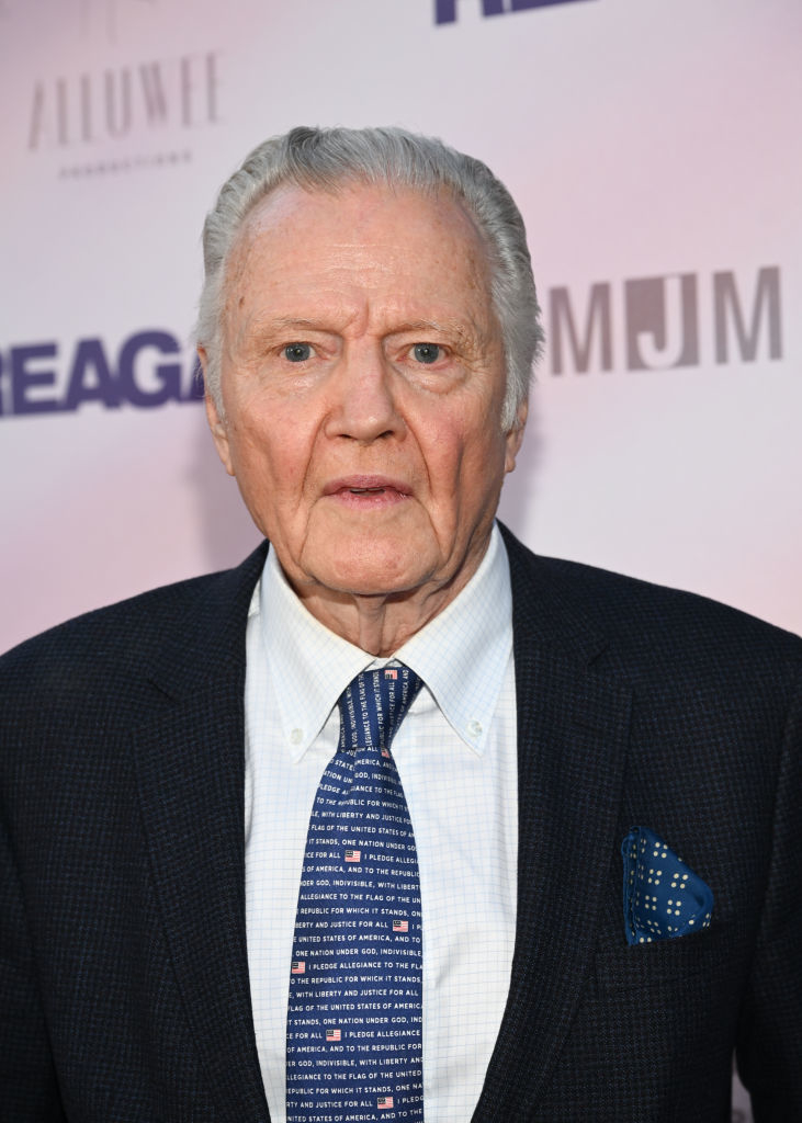 Jon Voight at a red carpet event, wearing a dark suit with a white dress shirt and a blue tie with printed words