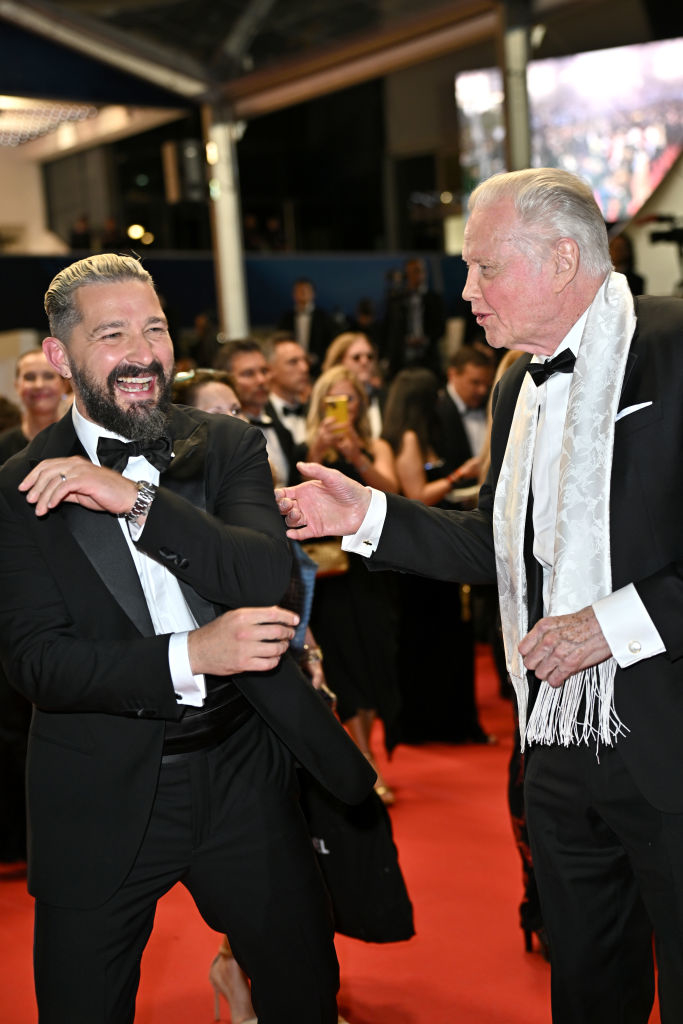Two unidentified men on the red carpet in tuxedos, one adjusting his sleeve, the other with a white scarf, in a lively conversation