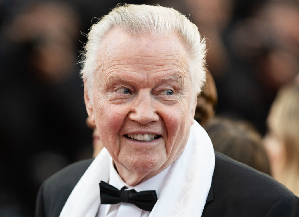 Jon Voight on the red carpet, wearing a black tuxedo with a bow tie and a white scarf. He is smiling and looking to his side