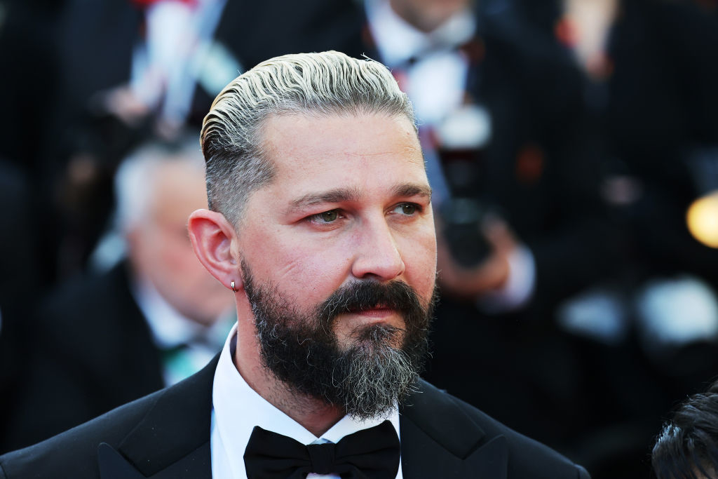 Shia LaBeouf on the red carpet wearing a black tuxedo and bow tie, with slicked-back hair and a full beard