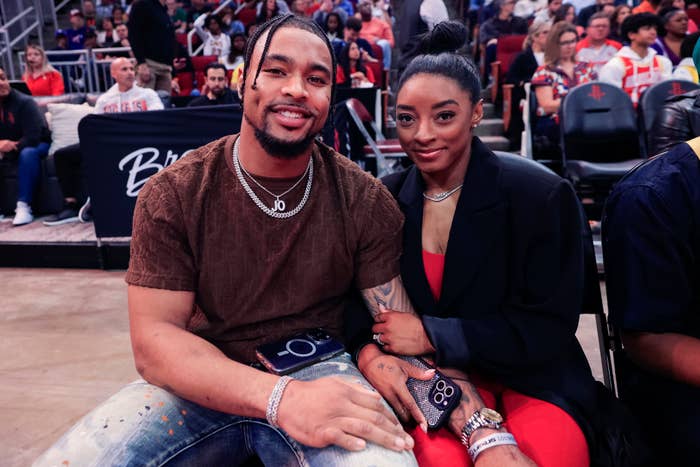 Simone Biles and Jonathan Owens sit courtside at a basketball game. Jonathan wears a dark t-shirt and necklaces, while Simone wears a blazer over bright outfit