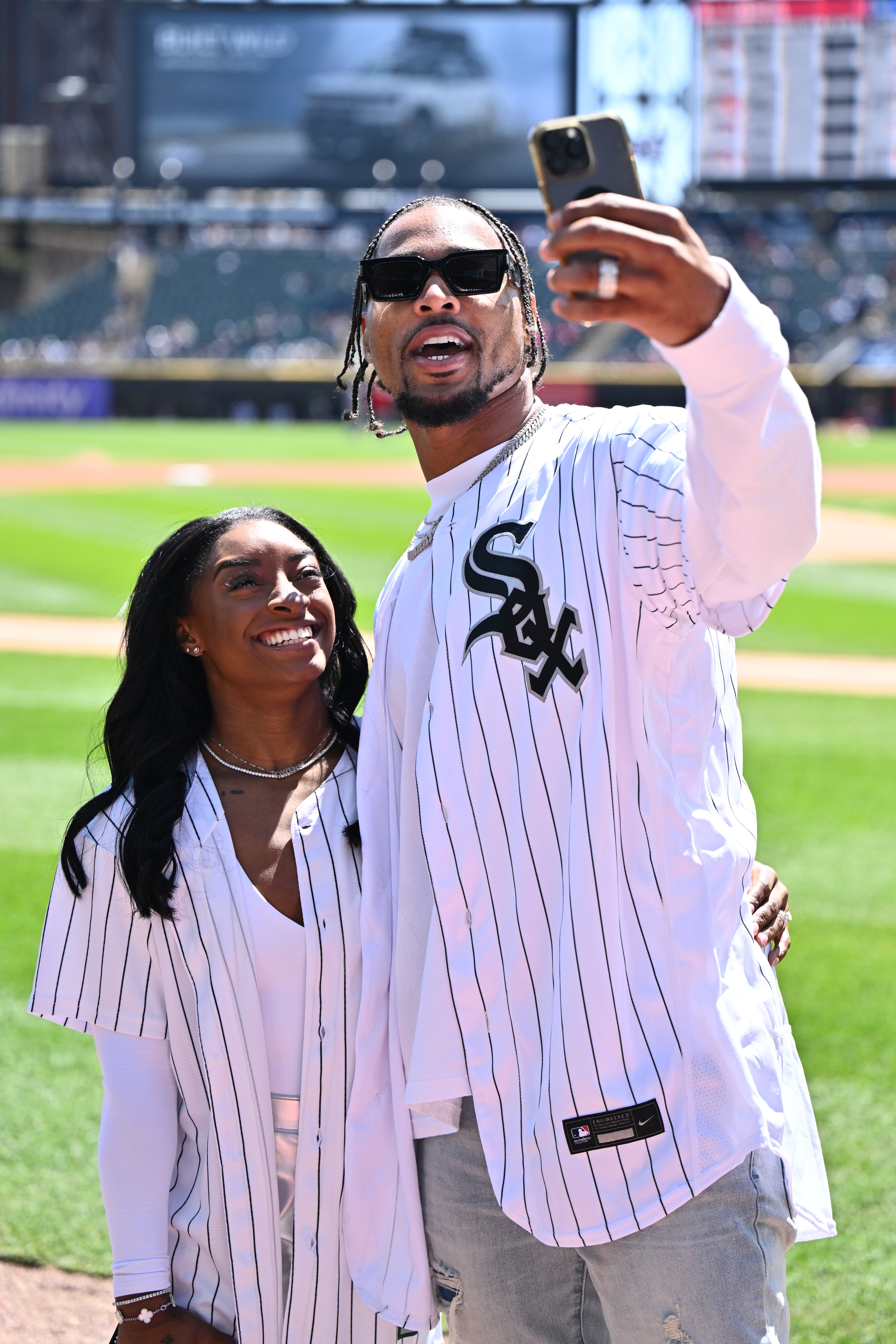 Simone Biles and Jonathan Owens take a selfie on a baseball field. They both wear matching white pinstriped Chicago White Sox jerseys