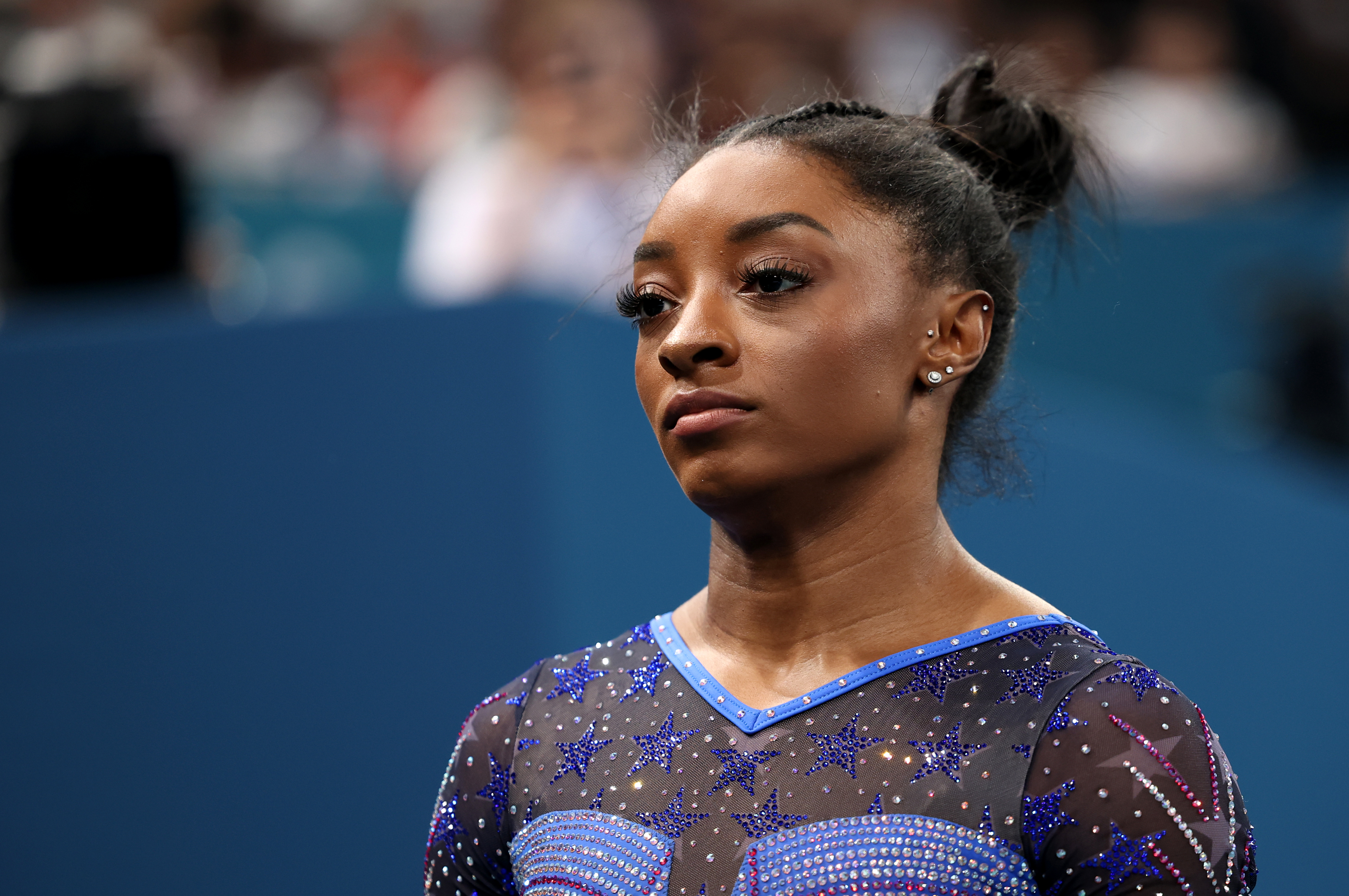 Simone Biles stands in a sparkling leotard at a gymnastics competition, looking focused and determined