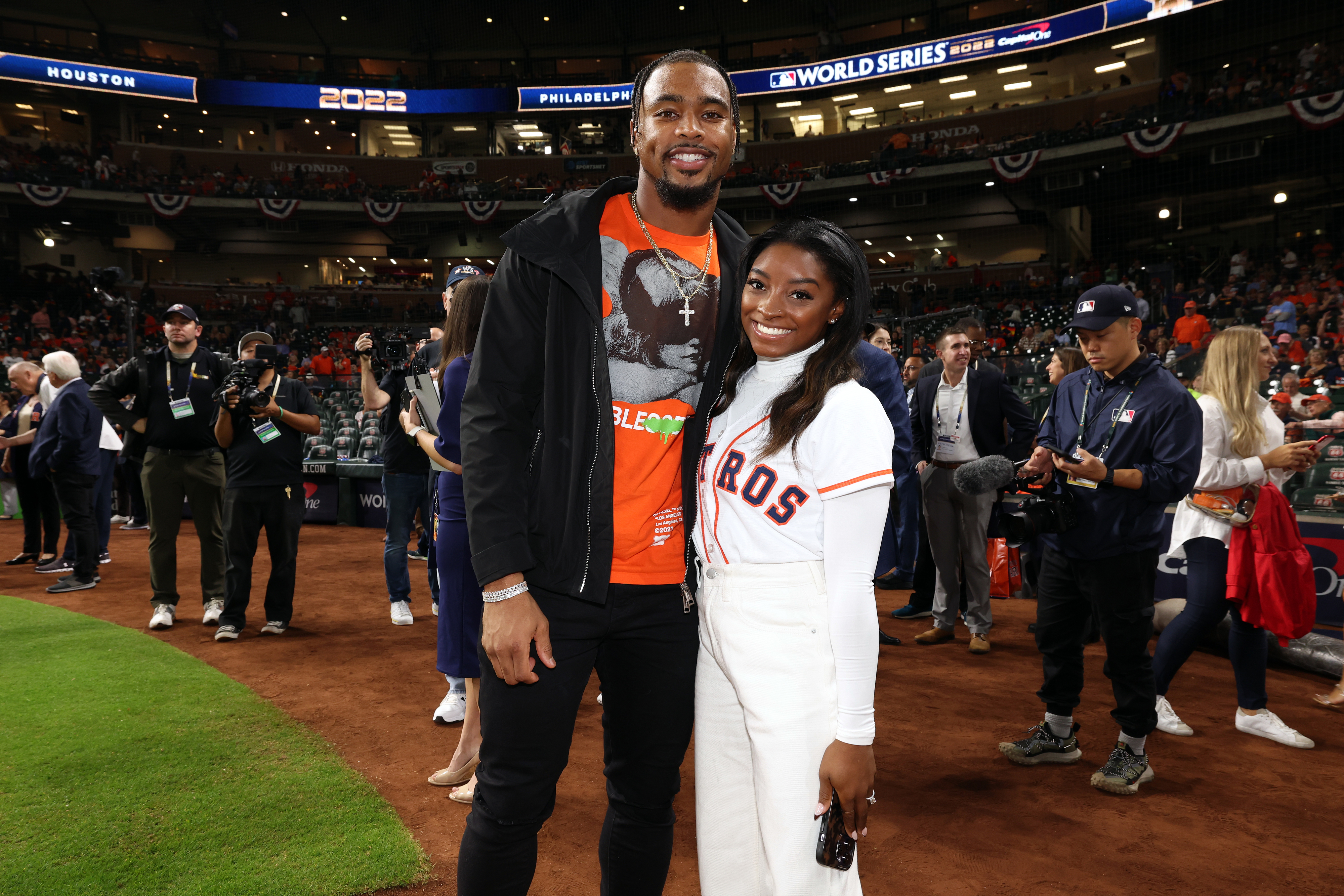 Jonathan Owens and Simone Biles smiling together on a baseball field during a game, Simone wearing an Astros jersey. People and event ambience in the background