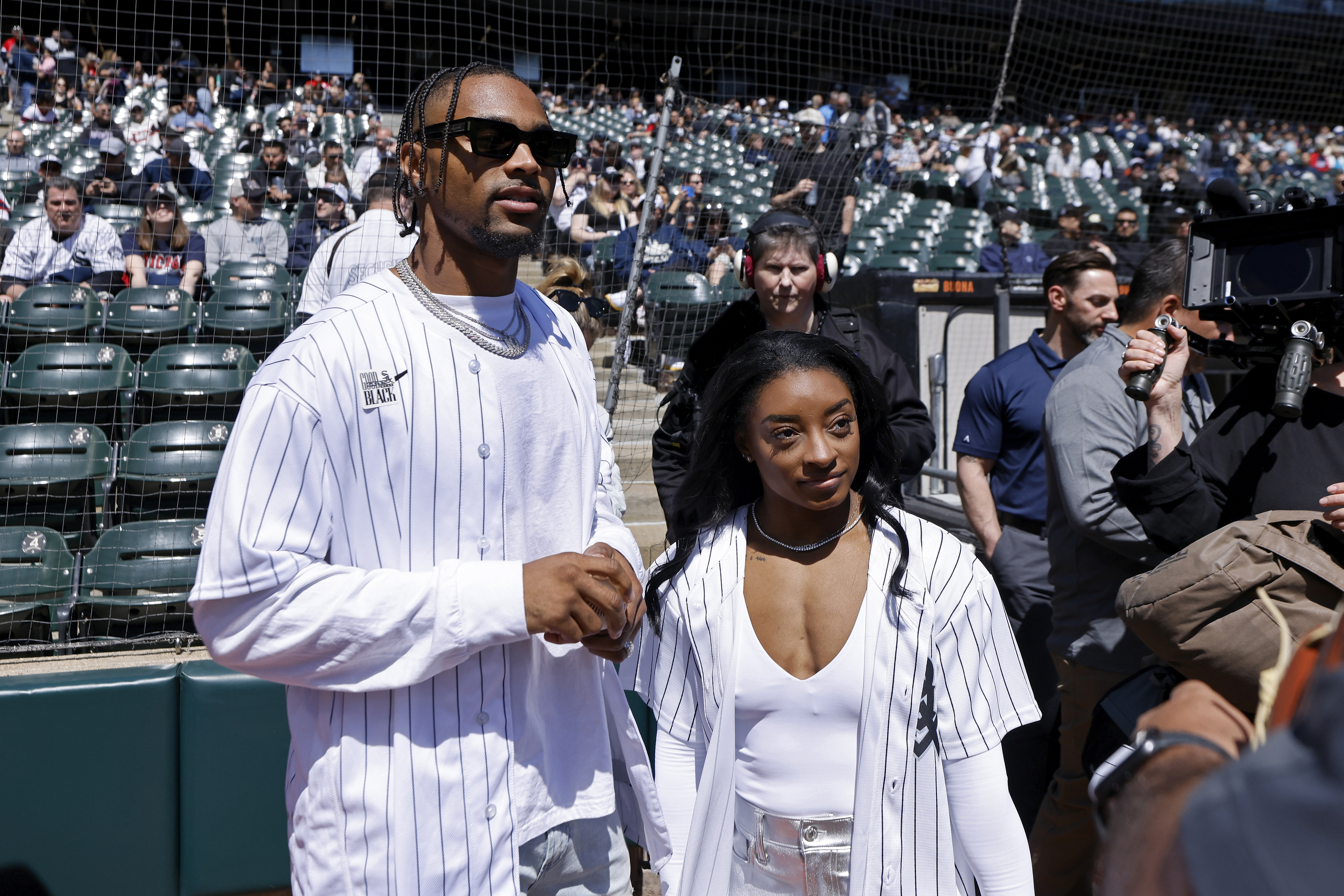 Jonathan Owens and Simone Biles stand together at a baseball game, wearing matching white jerseys, with spectators in the background
