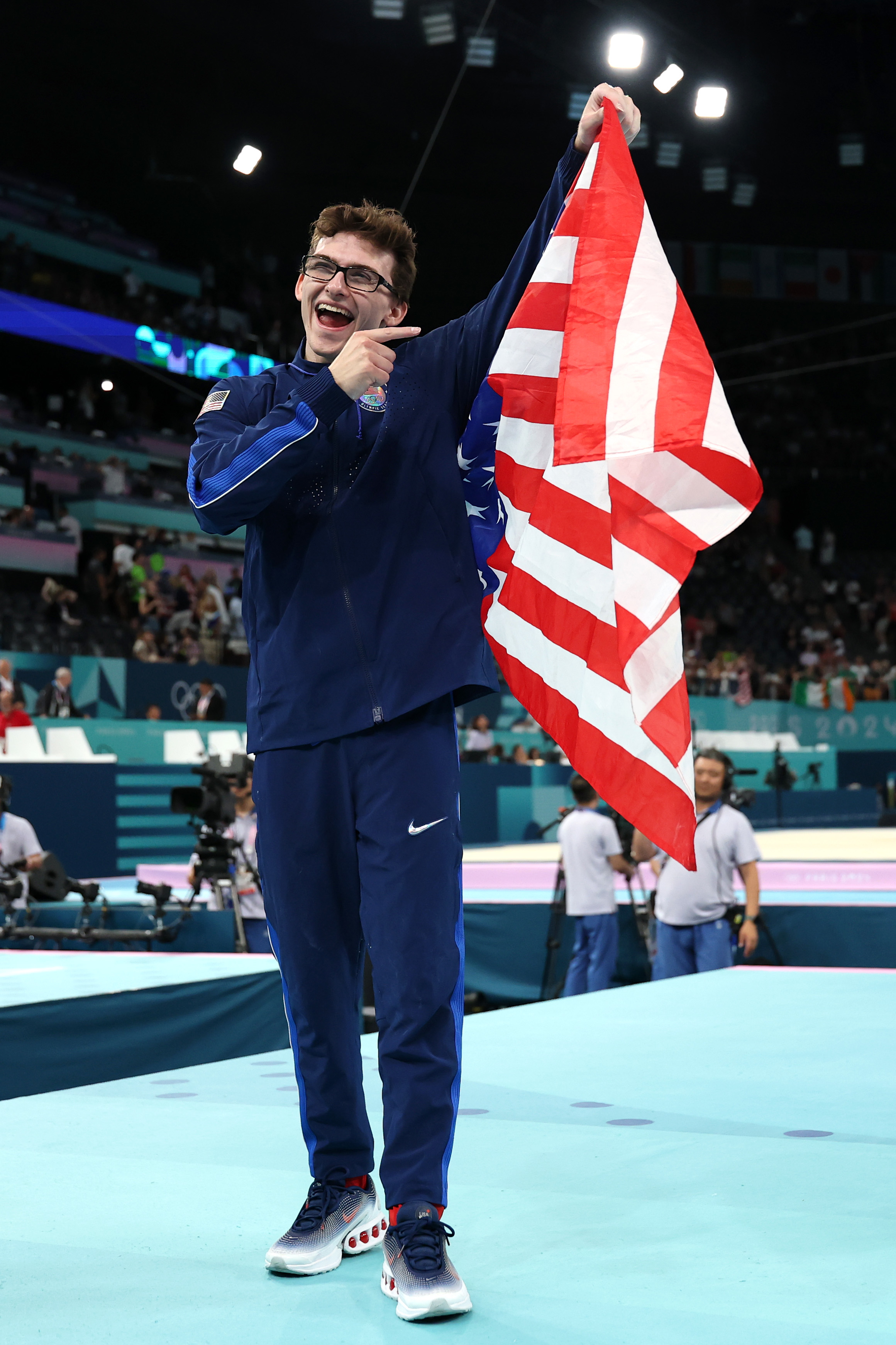 A person dressed in athletic wear holds up an American flag while smiling and pointing to it at a sporting event