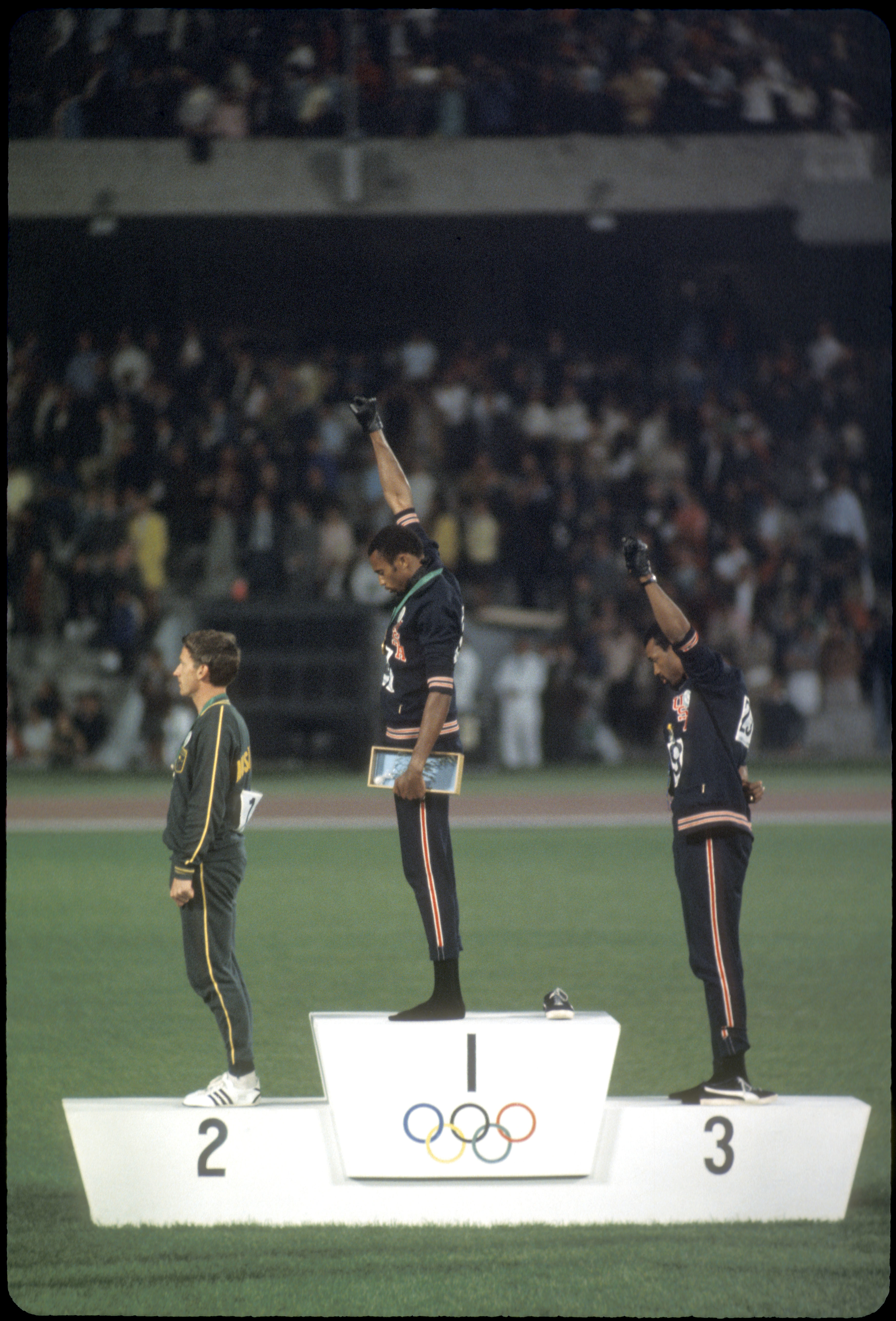 Tommie Smith and John Carlos stand on the Olympic podium with raised fists, while Peter Norman stands next to them