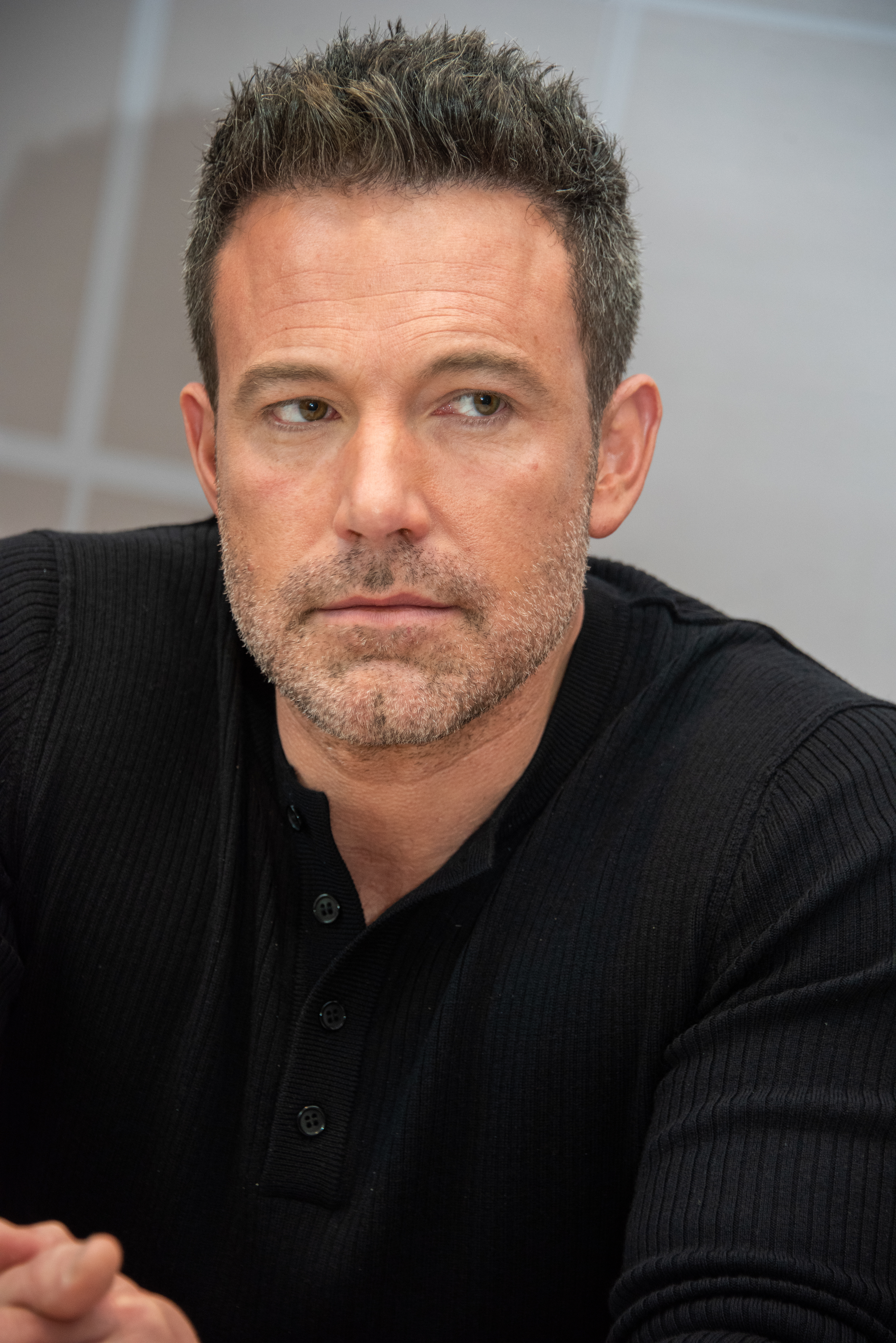 Ben Affleck in a black ribbed button-up shirt, looking thoughtful with a slight beard and short, styled hair
