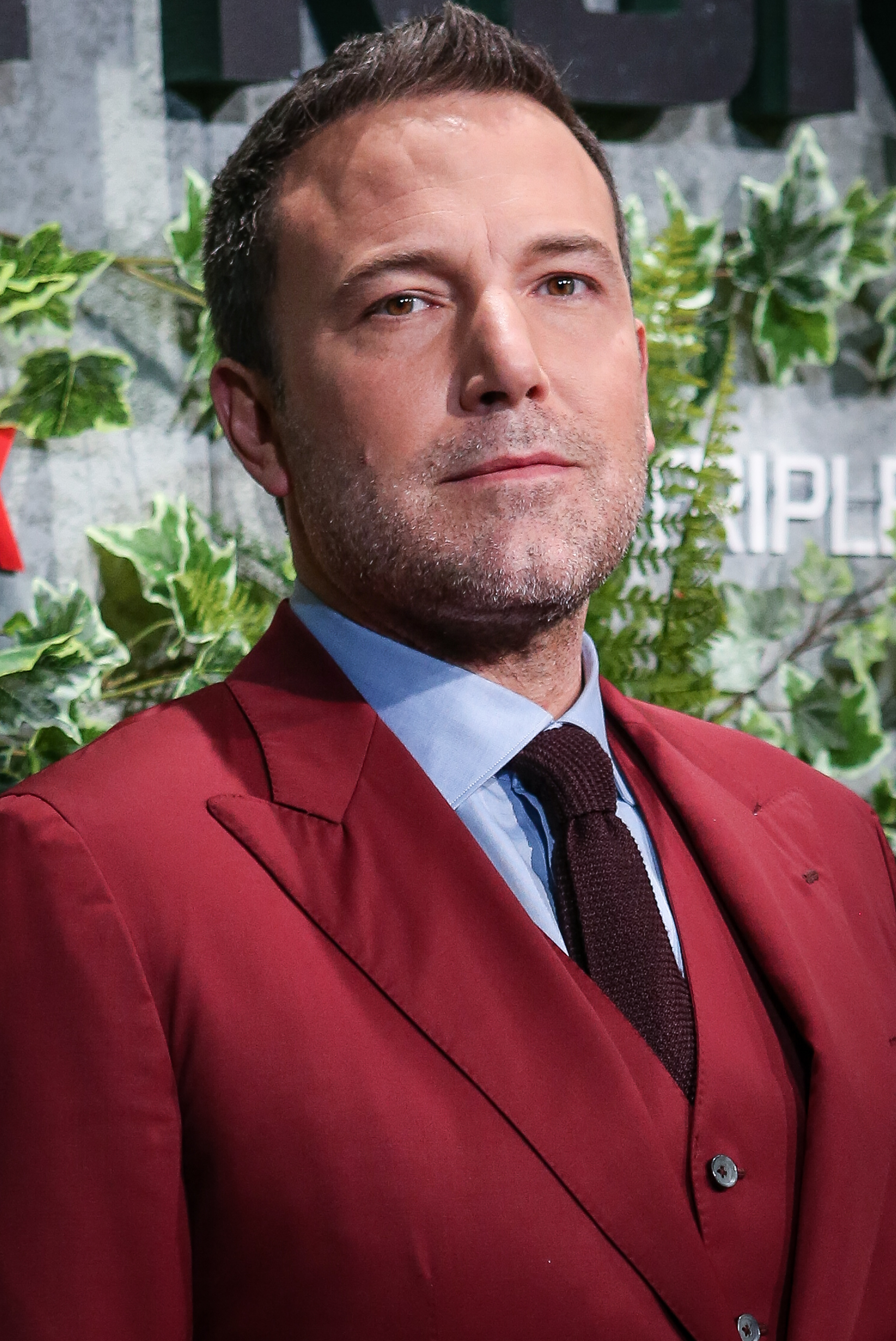 Ben Affleck attending a red carpet event in a stylish, dark red suit with a matching tie. The background includes green foliage and event branding