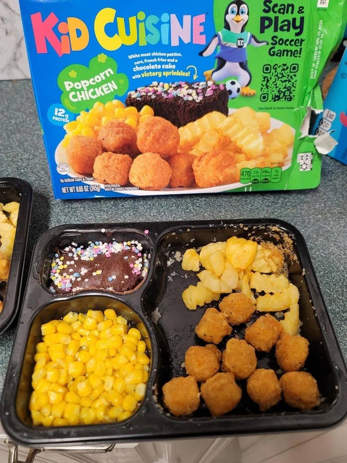 A Kid Cuisine tray with popcorn chicken, fries, corn, and a chocolate brownie with sprinkles. The meal box is also visible in the background