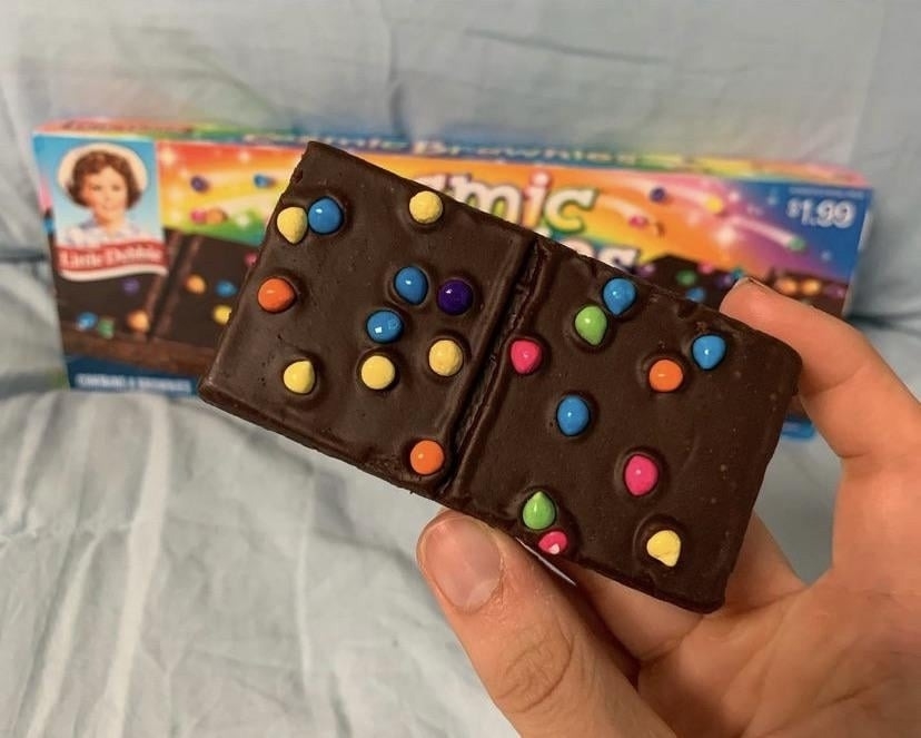 A hand holds a Little Debbie Cosmic Brownie, topped with colorful candy-coated chocolate pieces. The box in the background shows the product and price