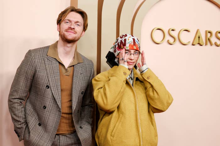 Finneas and Billie Eilish posing at the Oscars event. Finneas is in a plaid suit; Billie wears a yellow jacket and a headscarf with a pattern, adjusting her glasses