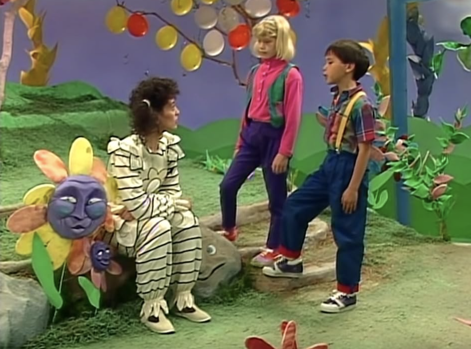 Three people talking in a whimsical, colorful set. The seated person is in a striped costume; the two others are children in casual clothing