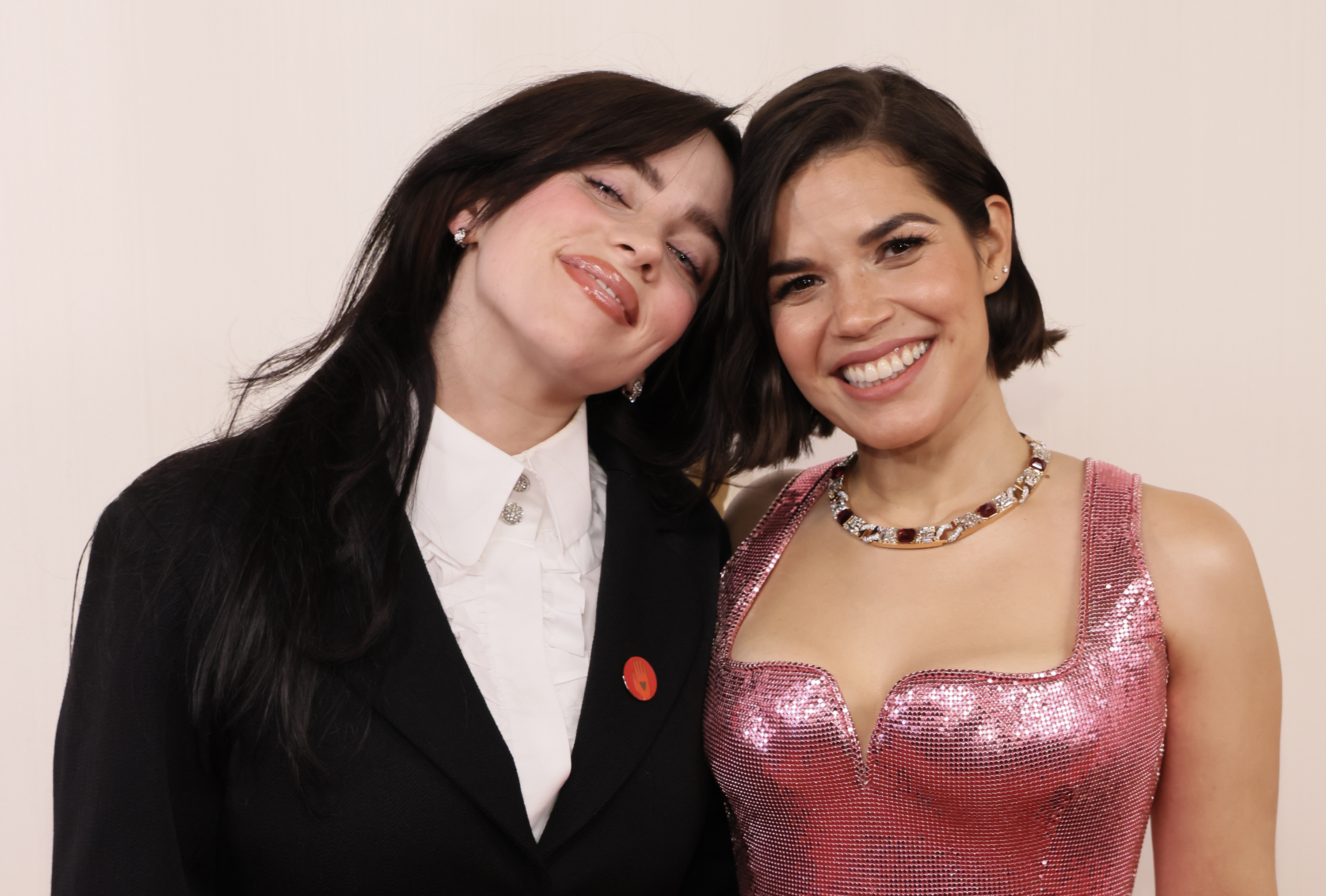 Billie Eilish in a chic suit and America Ferrera in a shimmering pink dress, smiling together for a photo