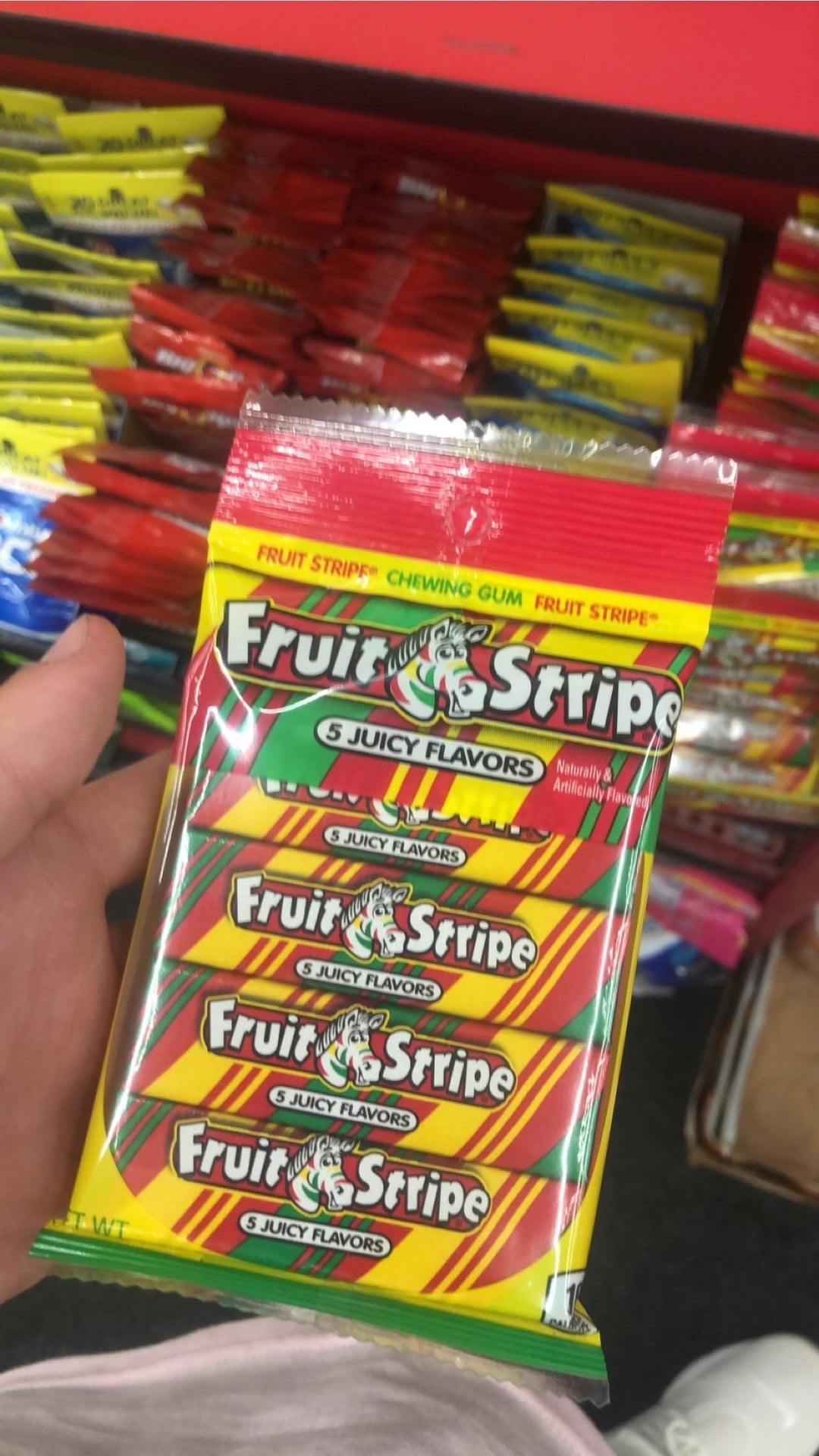 A hand holding a pack of Fruit Stripe gum featuring the zebra mascot, with text highlighting "5 Juicy Flavors" in a store aisle filled with more packs of gum
