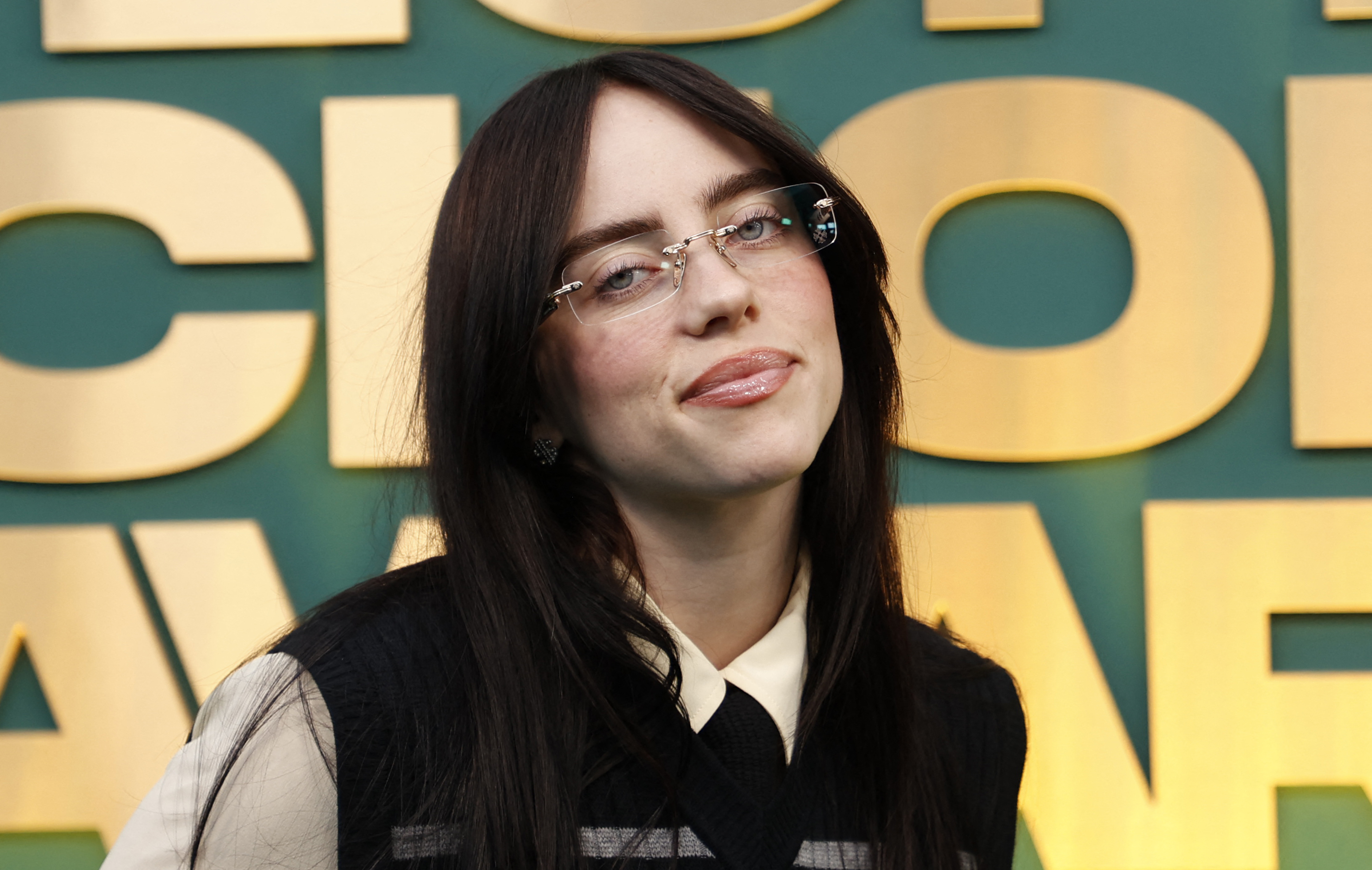 Billie Eilish at an event, wearing a black vest over a collared shirt and glasses, standing in front of a backdrop with large yellow lettering