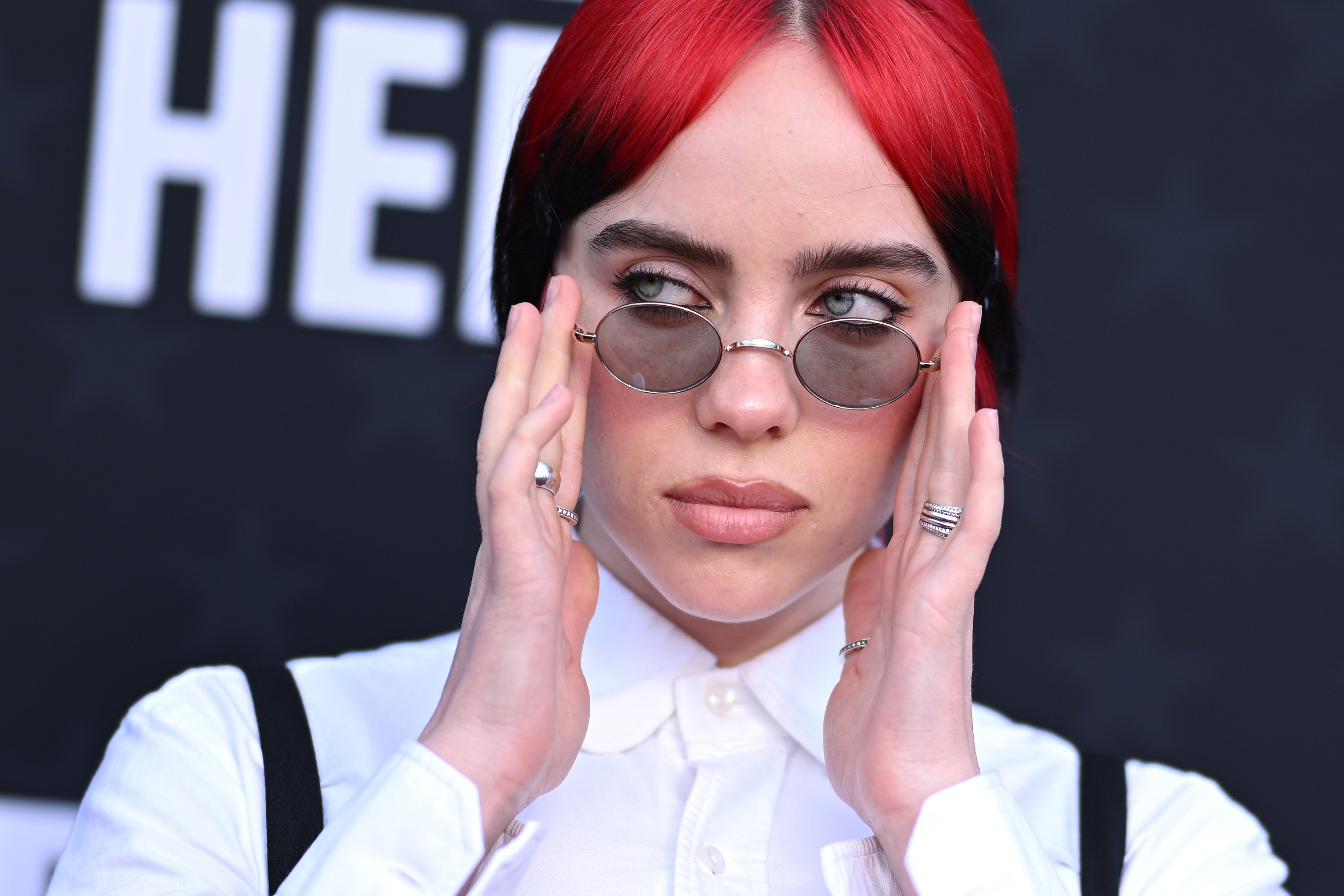 Billie Eilish at a red carpet event, wearing a white button-up shirt, round sunglasses, and holding her hands up to her face