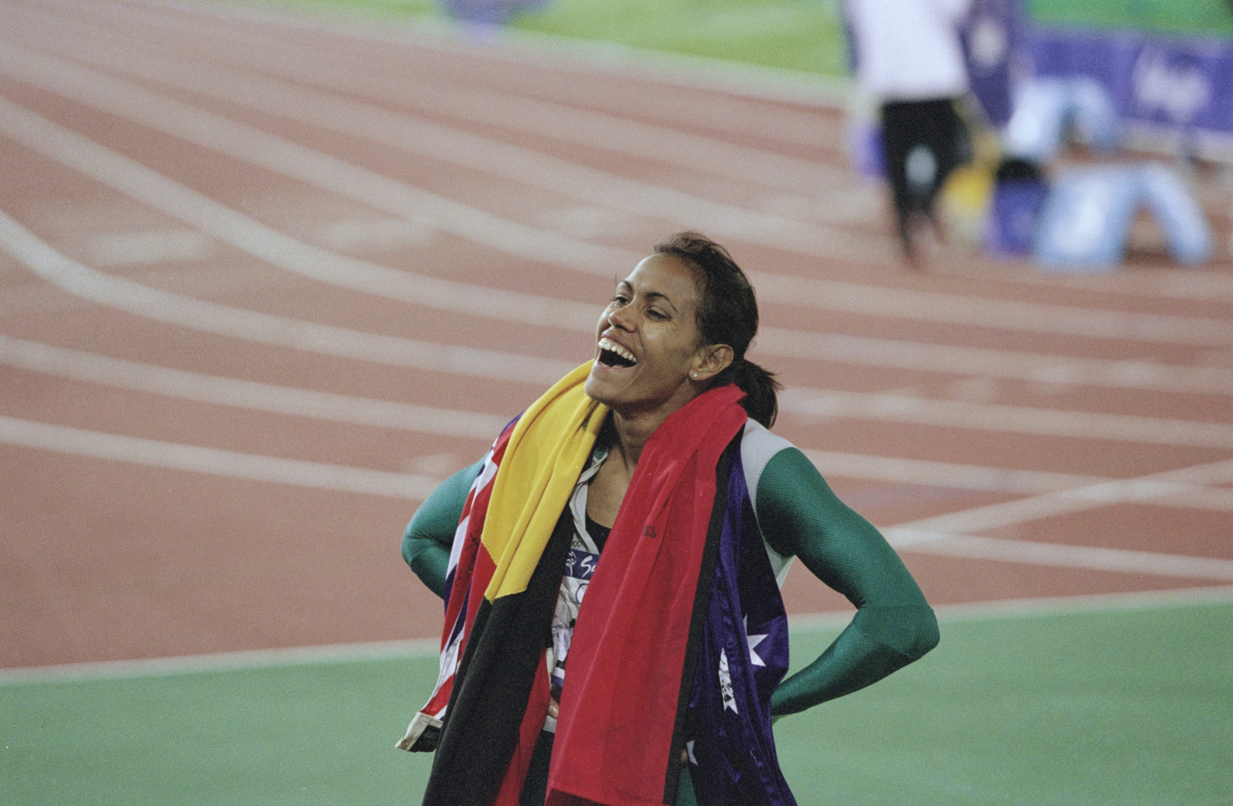 Female athlete standing on a track, smiling with hands on hips, draped in flags post-race