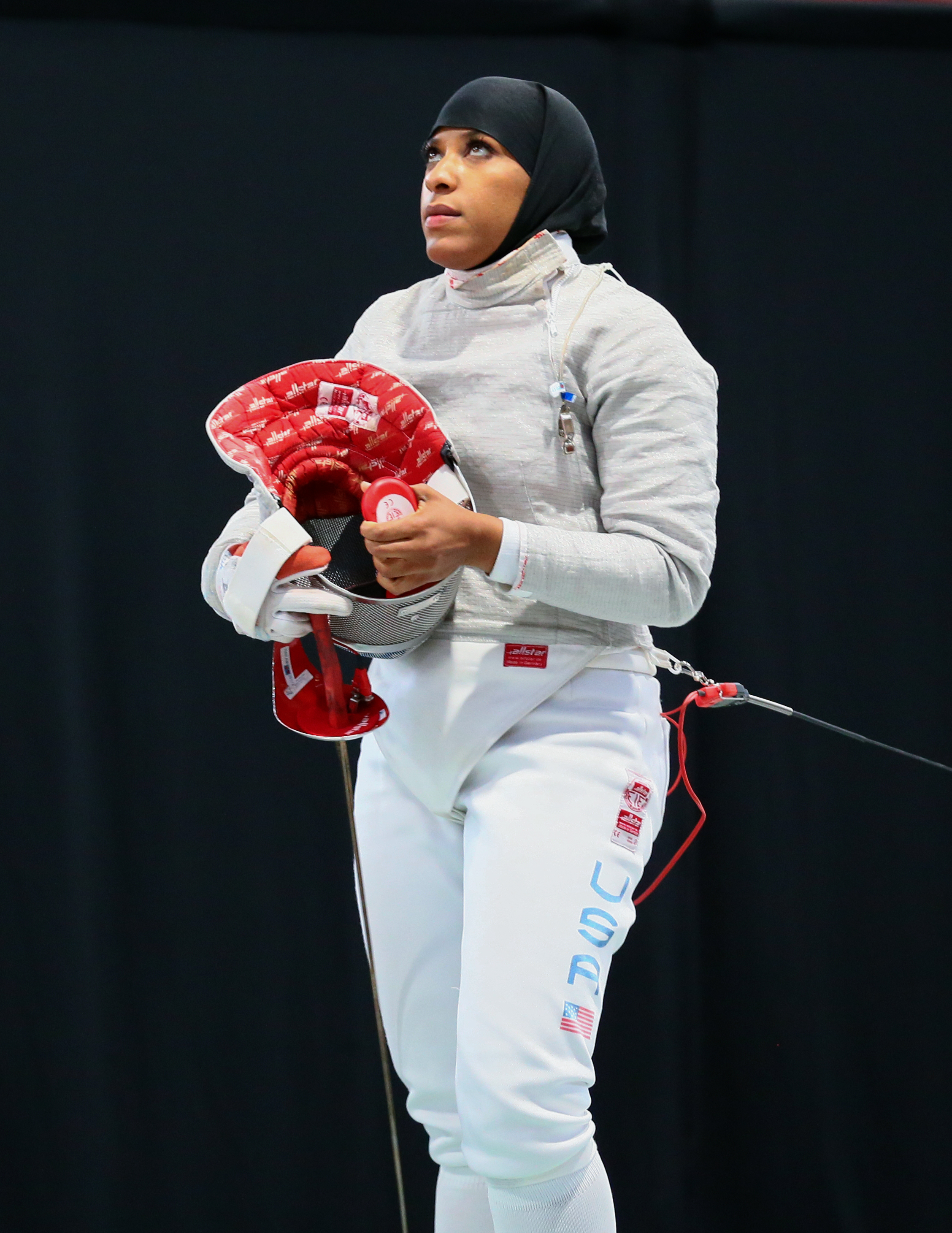 Ibtihaj Muhammad wearing fencing gear and holding her mask, standing on a fencing strip