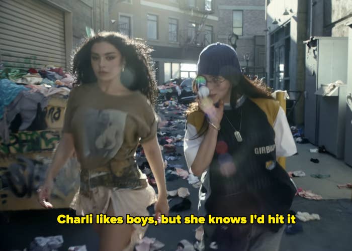 Charli XCX and Billie Eilish walk through an alley filled with scattered clothes; Charli wears a casual outfit, and Rina is dressed in a sports-casual ensemble