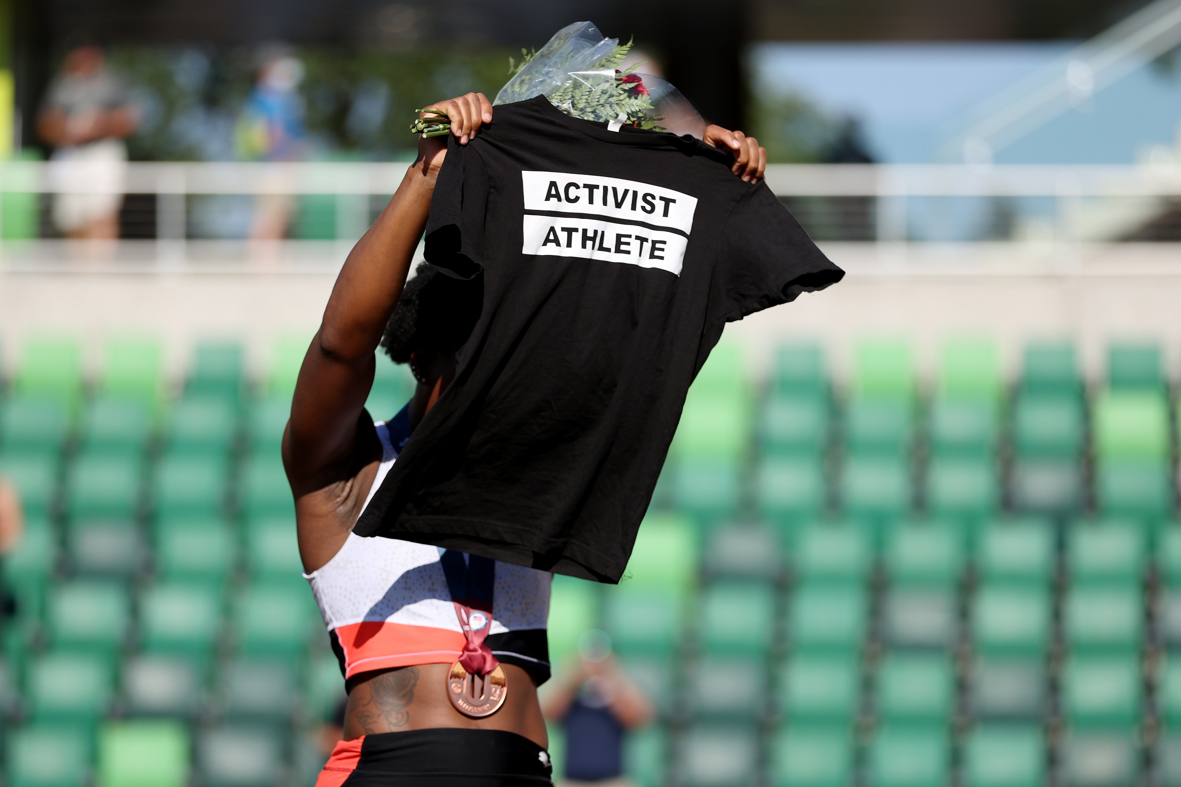 Athlete on podium holds up a shirt reading "Activist Athlete" with a bouquet, obscuring their face, at a sports event