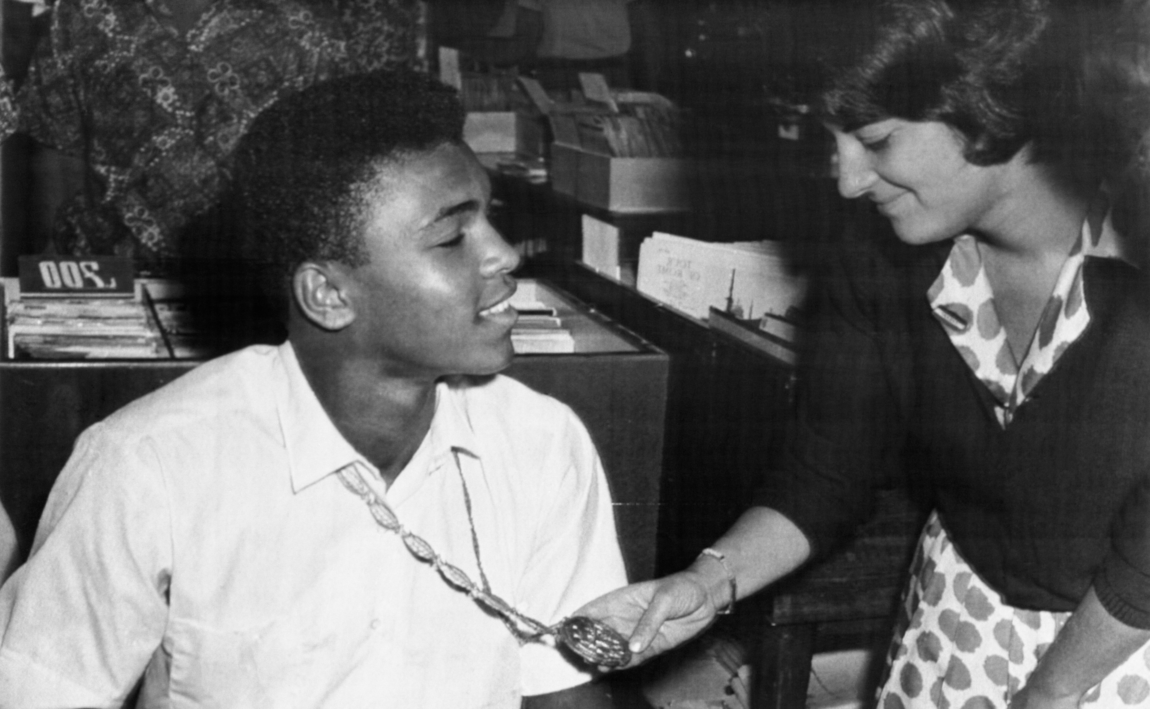 A young Muhammad Ali is seated, smiling, as a woman standing next to holds up the medallion around his neck