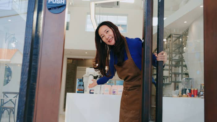 A smiling woman wearing a brown apron and blue shirt stands at the entrance of a modern bakery, holding the door open