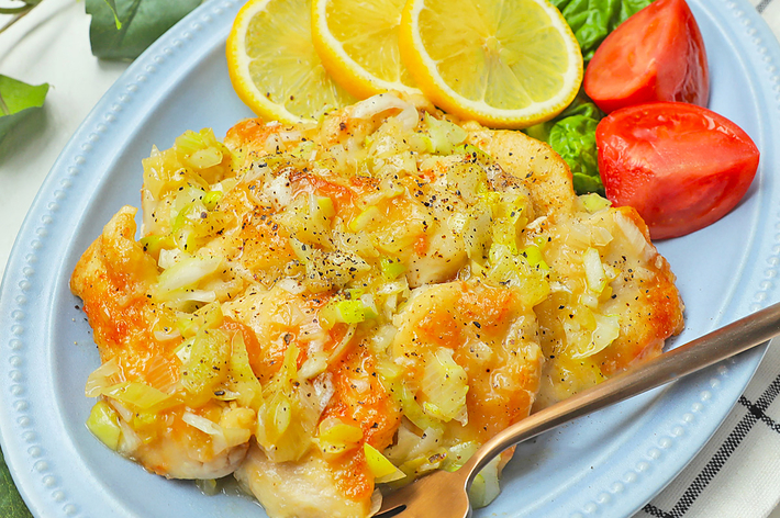 白い皿の上にレモンスライスとトマト片が添えられたチキン料理。 フォークが横に置かれ、背景に緑の葉が見える。
