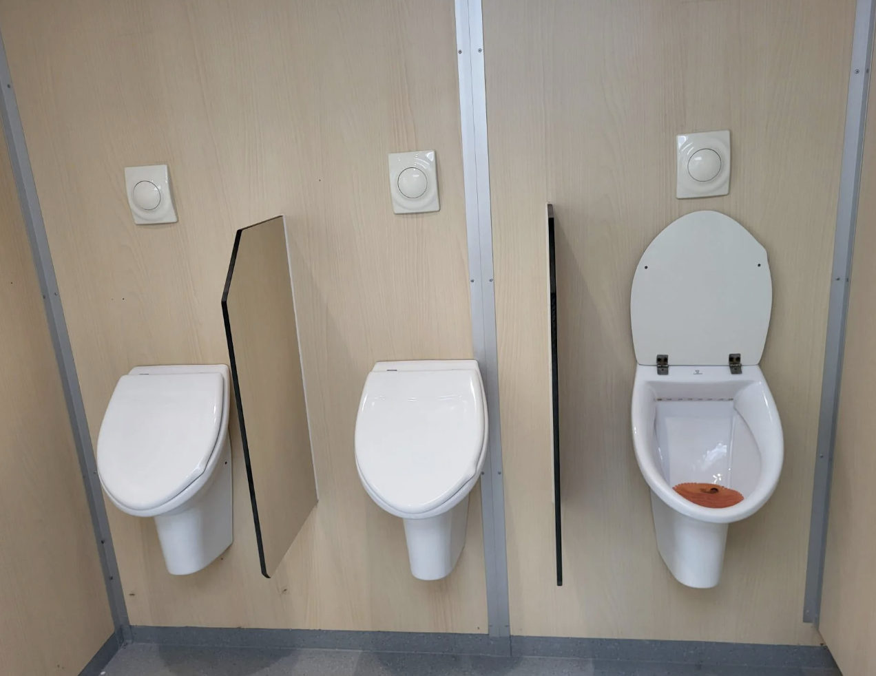 Three restroom urinals mounted on a beige wall, with the rightmost urinal appearing dirty