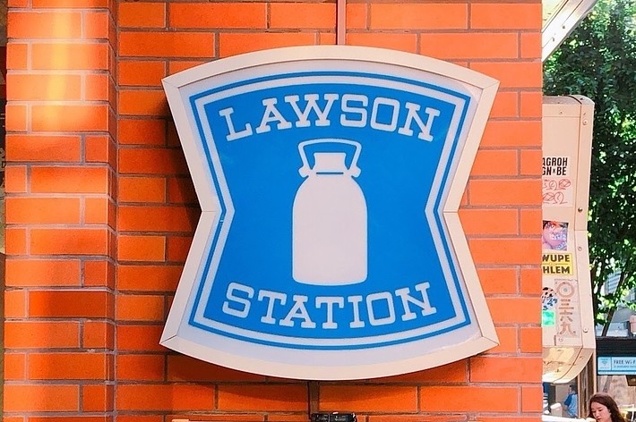 白い牛乳瓶のアイコンが描かれた「LAWSON STATION」の看板と、その下に「OPEN 24 HOURS」と表示された標識。