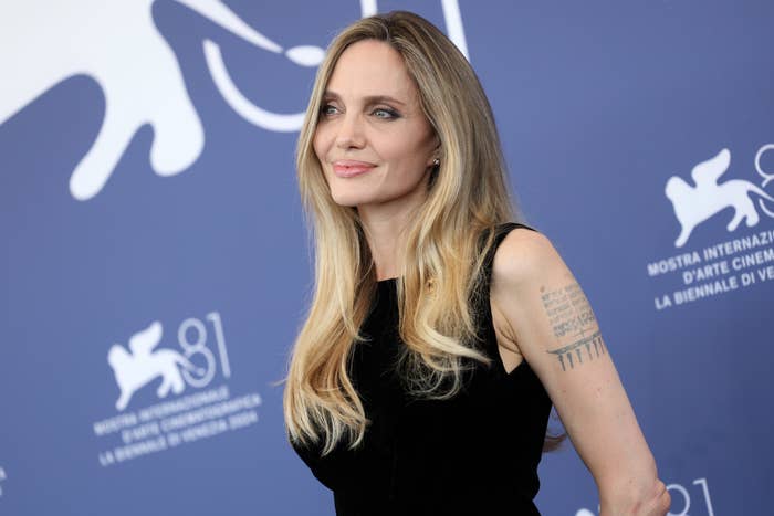 Angelina Jolie at a film festival, wearing a sleeveless dress, with long hair down, and tattoos visible on her right arm