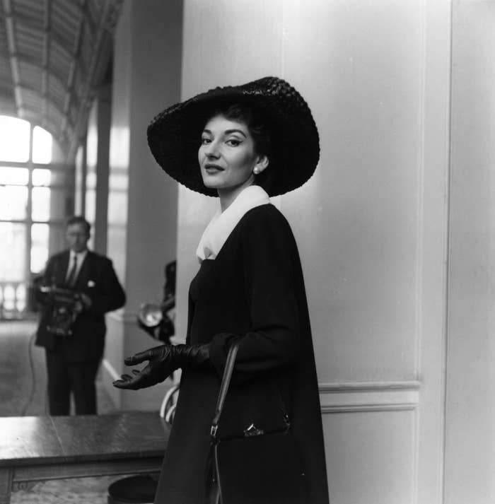 Maria Callas elegantly poses in a chic outfit with a wide-brim hat, gloves, and a handbag in an indoor setting