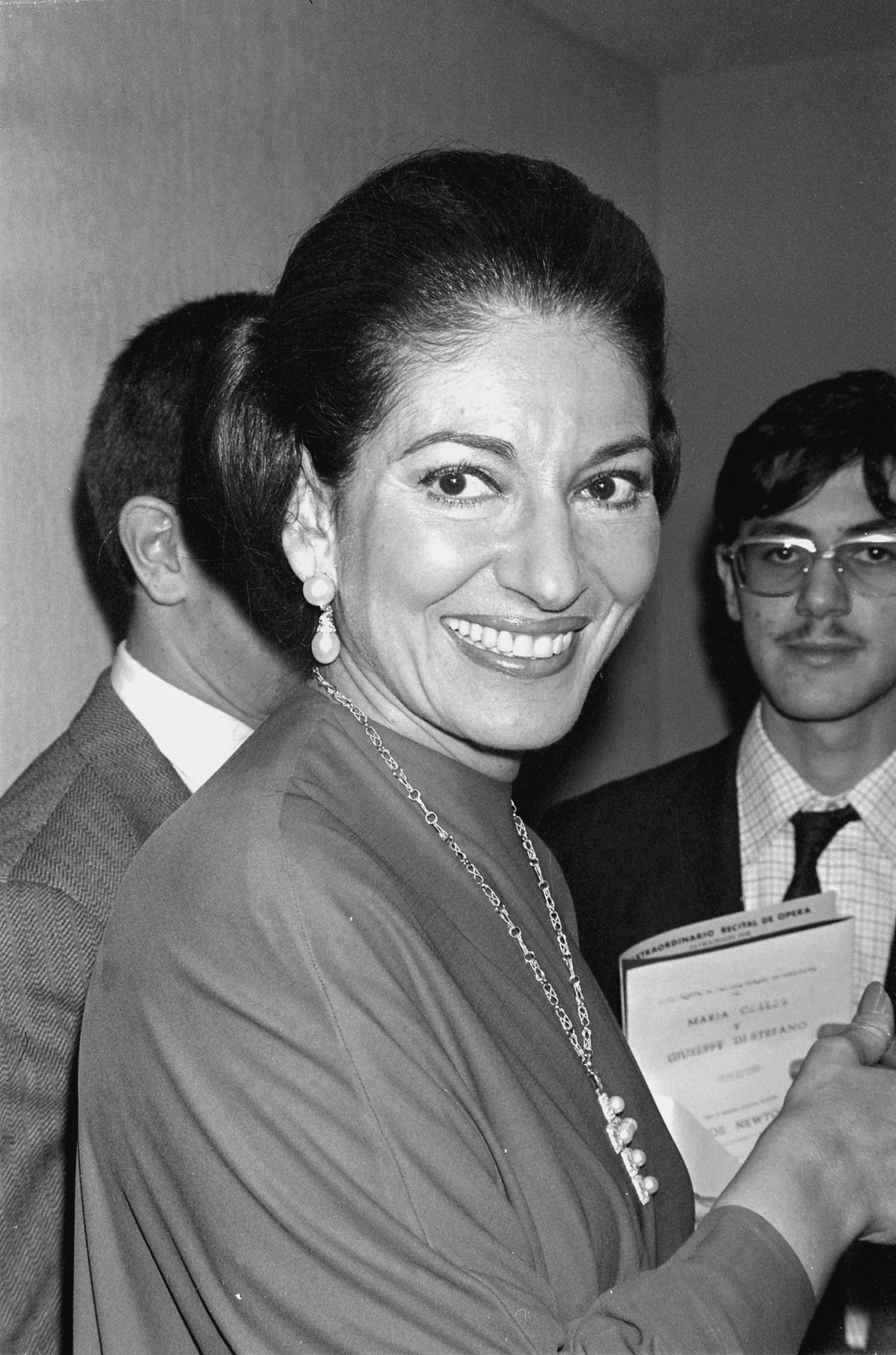 Maria Callas smiling and holding a program at an event with two men in the background. She is wearing a long dress, prominent earrings, and a necklace
