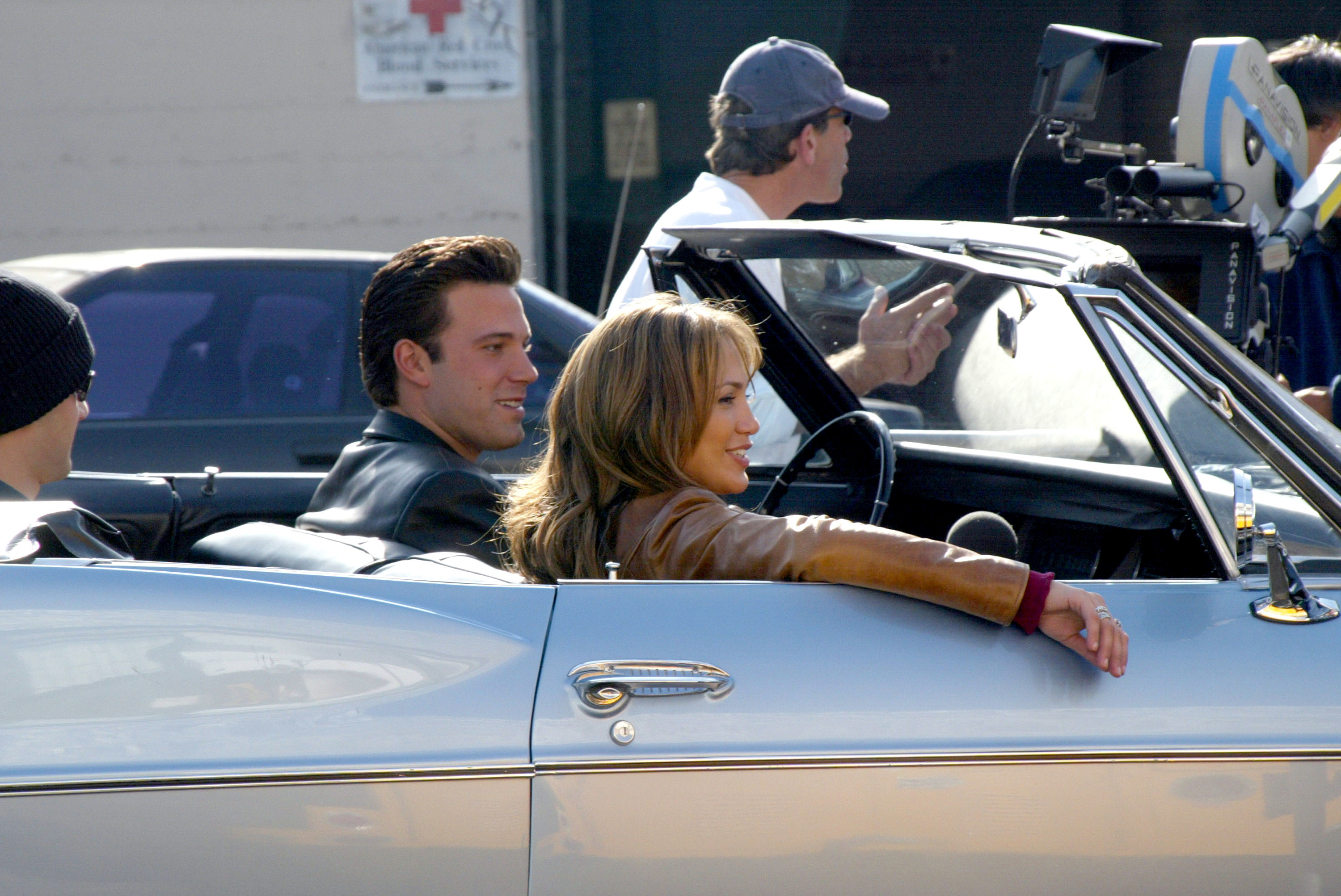 Ben Affleck in a leather jacket and Jennifer Lopez in a brown jacket sit in a convertible during a film shoot, surrounded by crew members and camera equipment