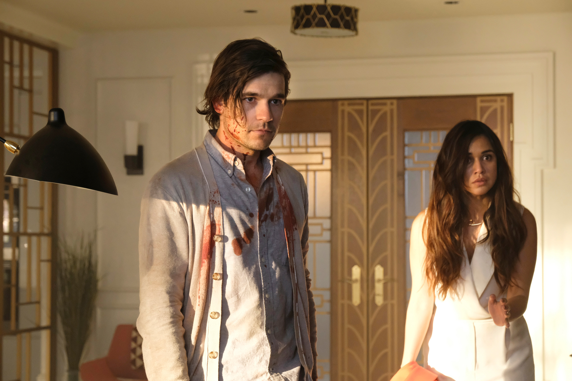 Jason Ralph, in a blood-stained shirt, stands next to Summer Bishil, who is gesturing while wearing a stylish dress, in an indoor setting