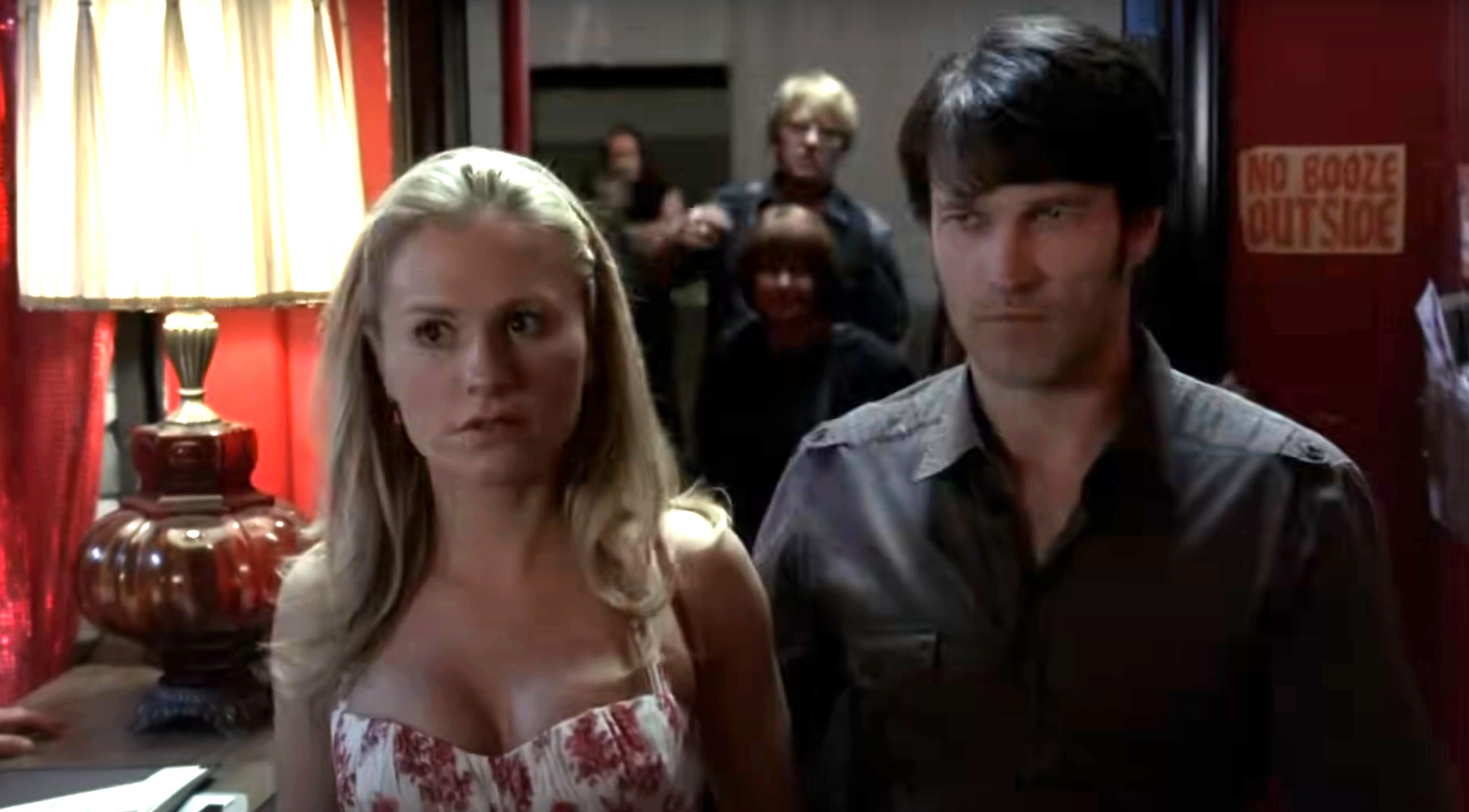 Anna Paquin and Stephen Moyer in a scene from a TV show, standing inside a dimly lit room with people and a sign in the background
